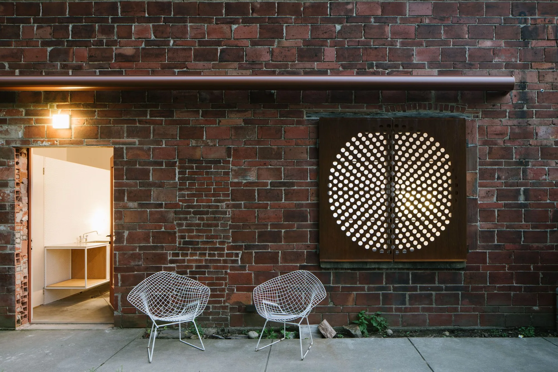 Two white wire chairs on a concrete patio in front of a red brick building with an open doorway and a circular perforated metal shutter.