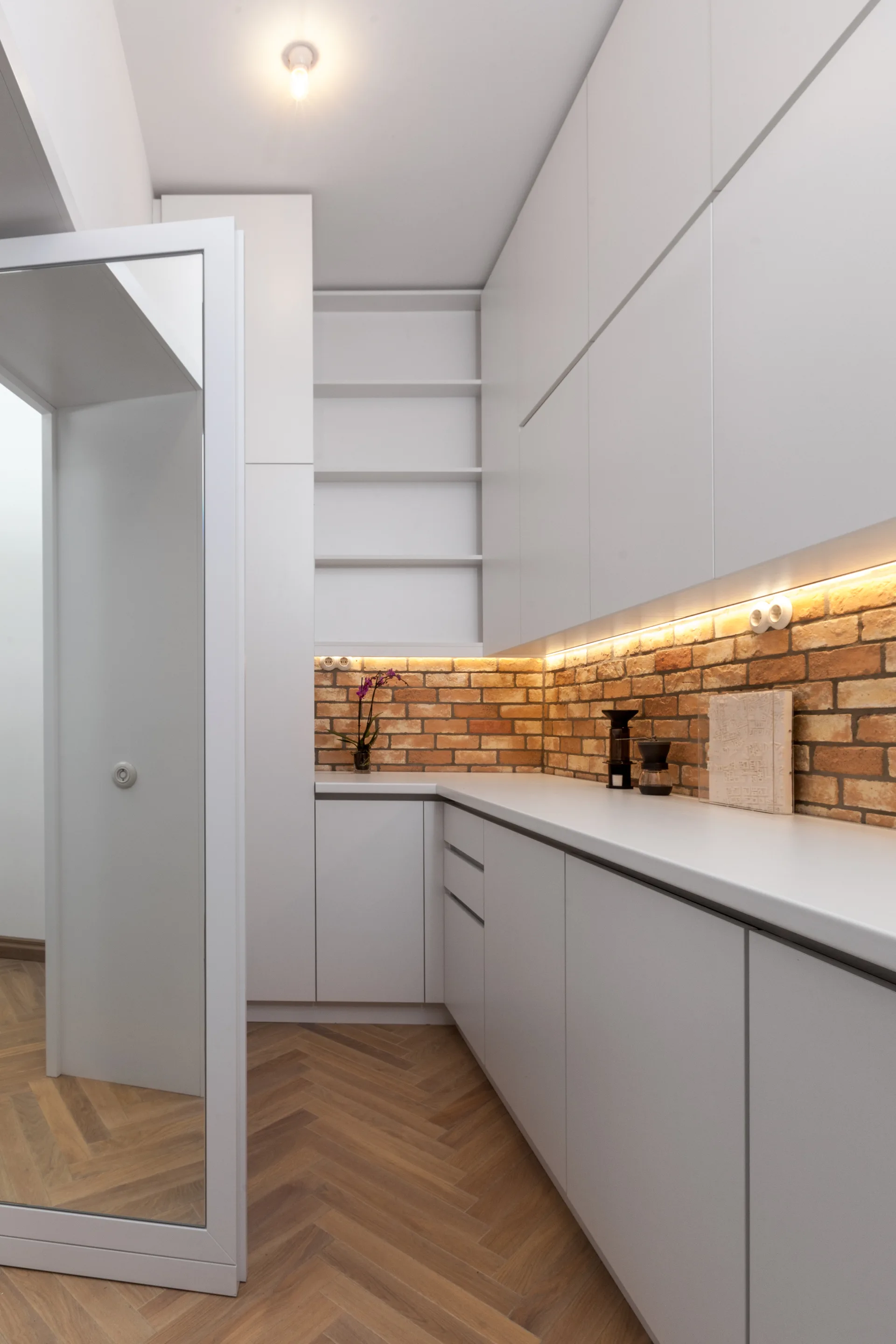 A modern kitchen featuring flat-panel white cabinets, an exposed brick backsplash with warm lighting, and a light wood herringbone floor.