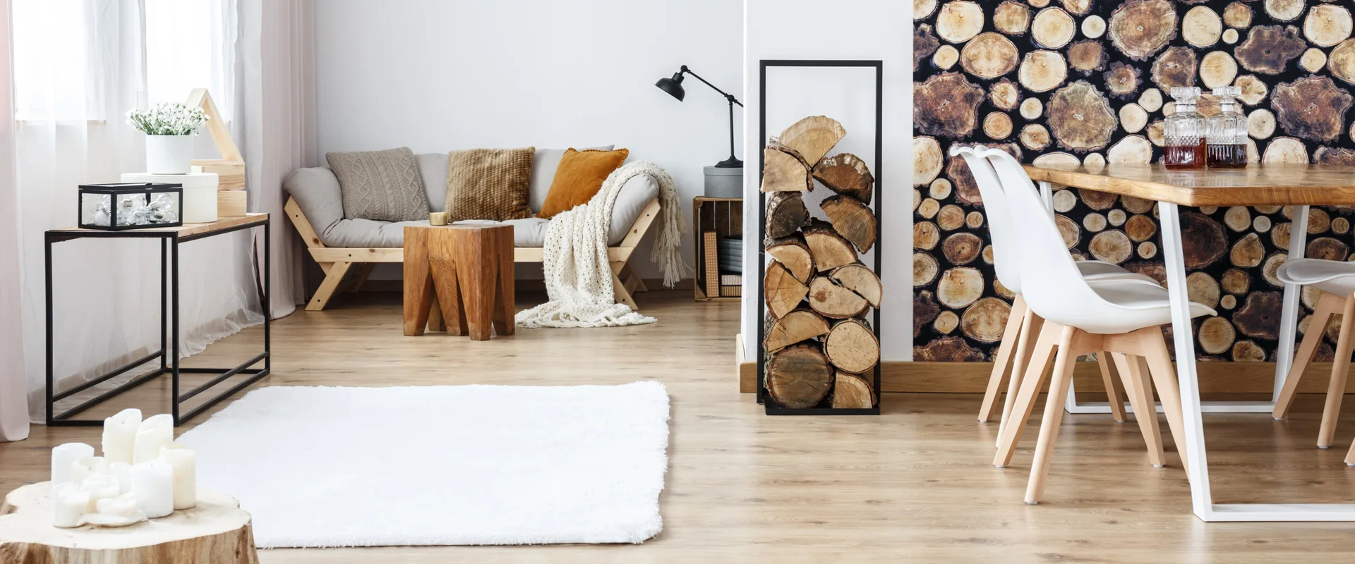 A bright open-concept room features a grey sofa with textured pillows, a black firewood rack, and a dining table set against a log-patterned accent wall.