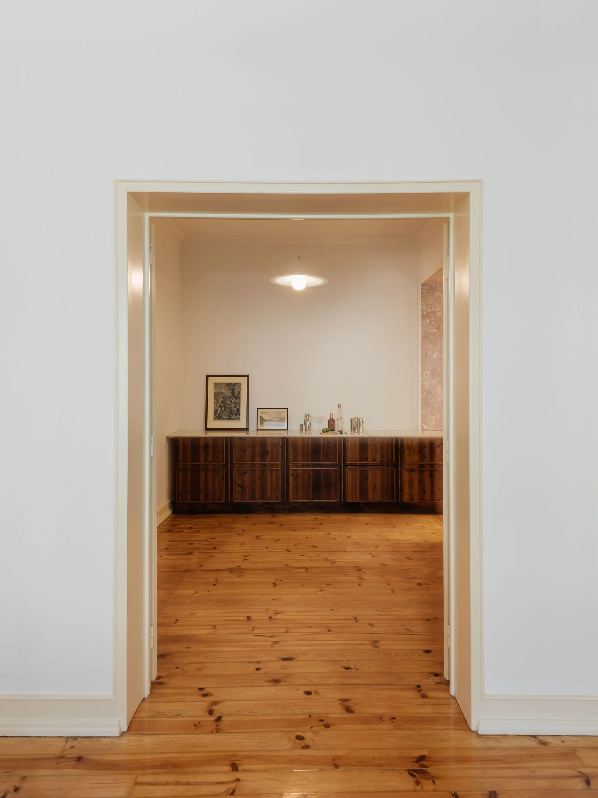 A view through a white door frame looking into a minimalist room featuring a long dark wood sideboard, pine floorboards, and a pendant light.