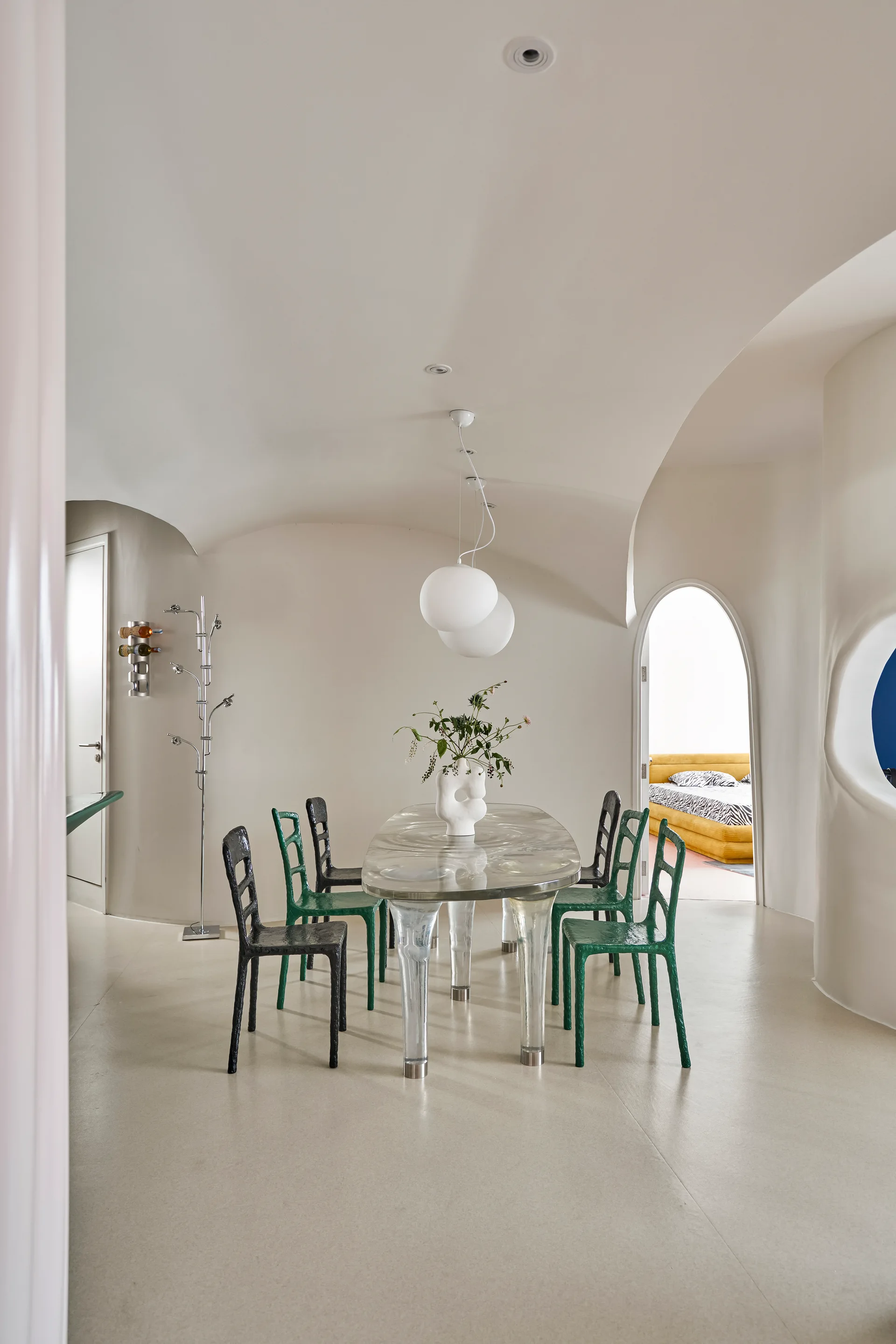 An off-white dining room features an oval clear resin dining table surrounded by green and black textured chairs under a curved plaster ceiling.