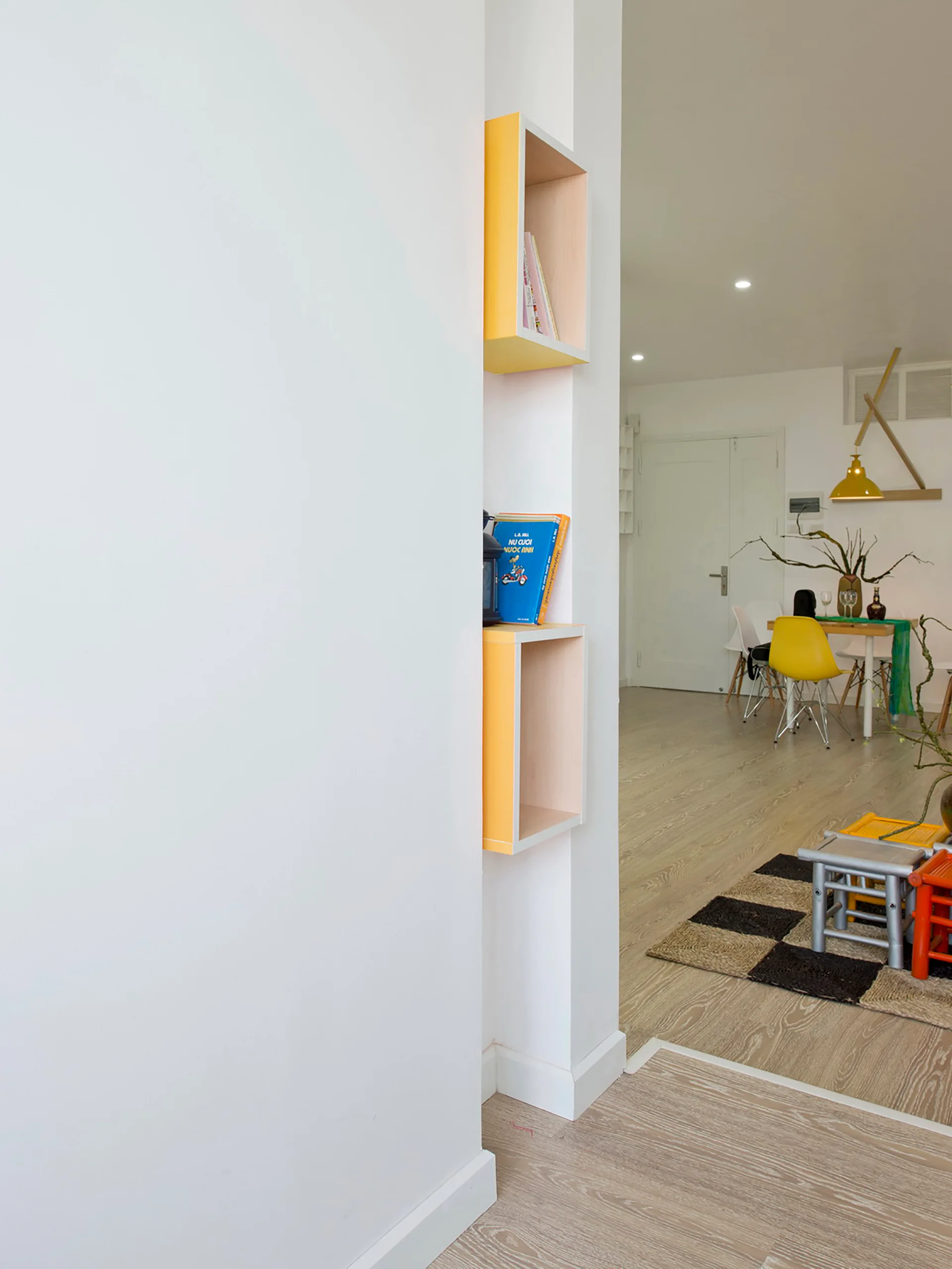 A view past yellow recessed wall shelves reveals a modern dining area with a wood table, yellow chairs, and a checkered rug.