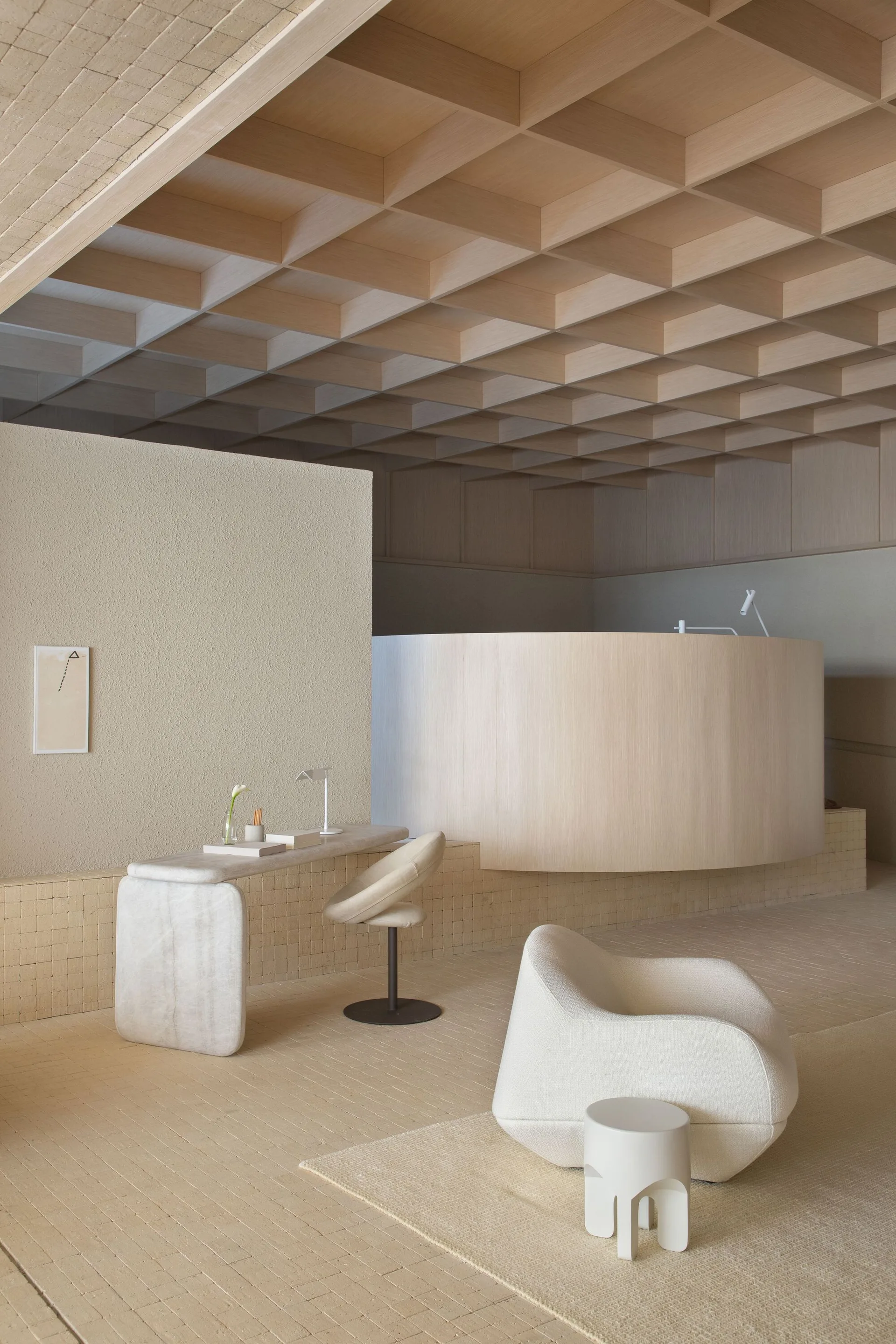 A minimalist room featuring a wooden coffered ceiling, a curved wood partition wall, a stone desk, and sculptural cream-colored armchairs.