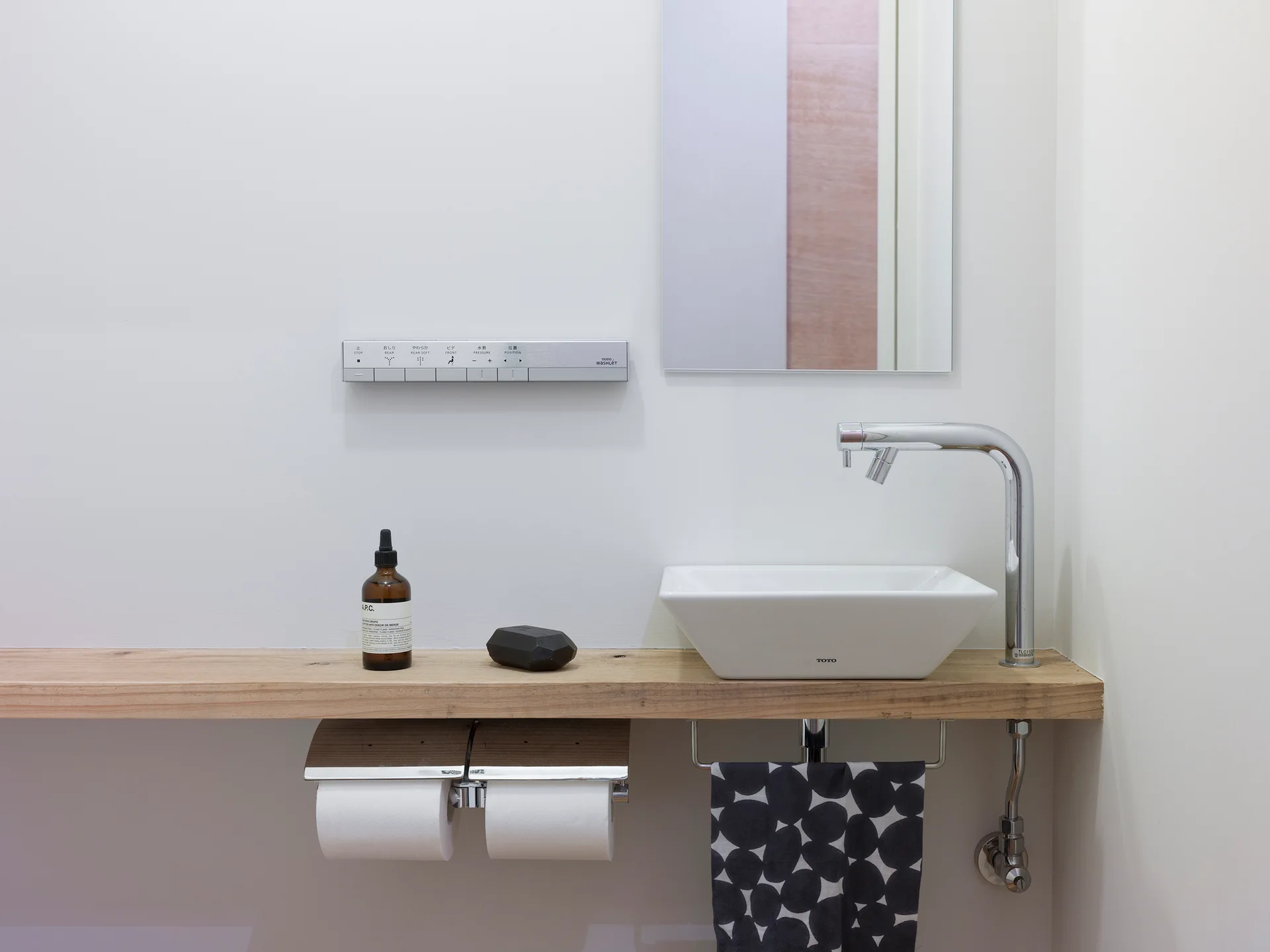 A minimalist bathroom vanity features a white vessel sink on a floating wood shelf beneath a wall-mounted electronic control panel and a rectangular mirror.