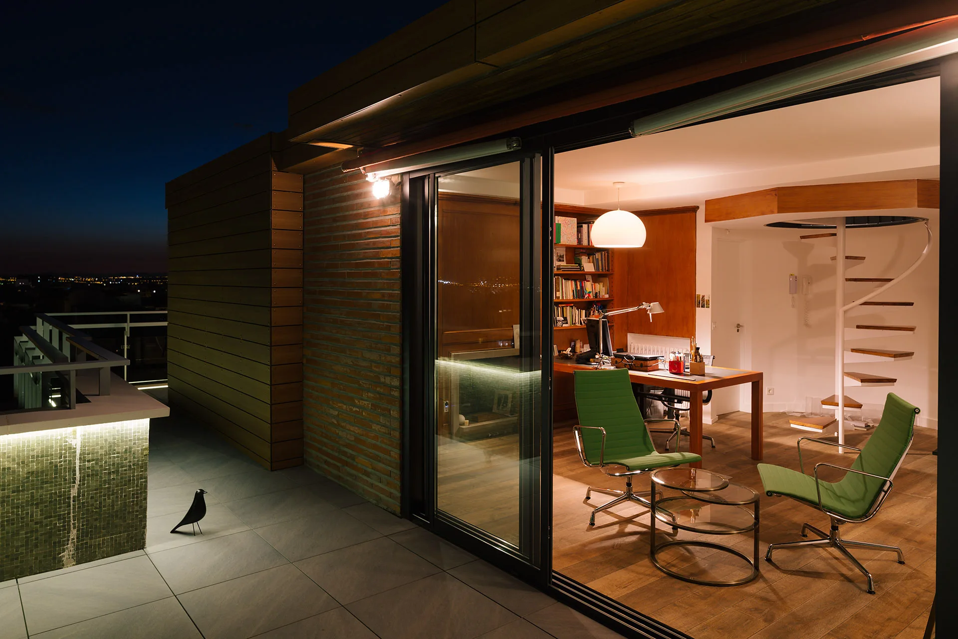 A warm home office with a wooden desk, green chairs, and a white spiral staircase seen through open glass sliding doors leading to a dark terrace.