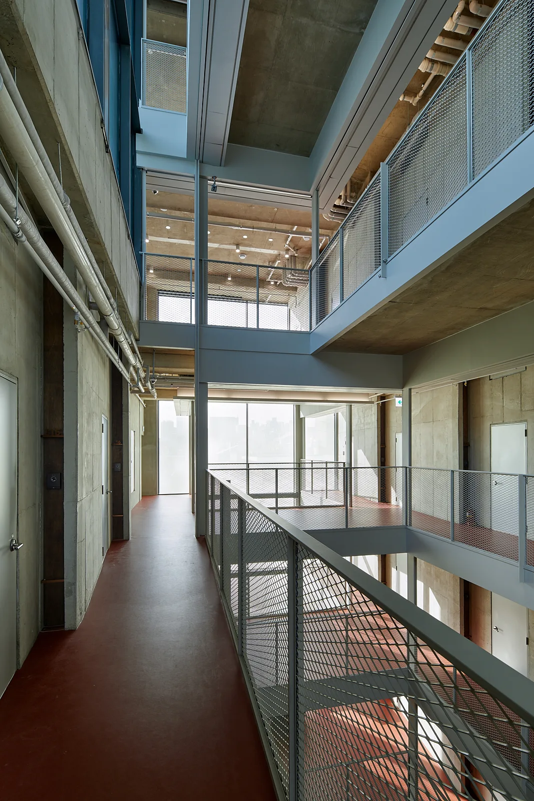 An industrial multi-level hallway with exposed concrete walls, steel mesh railings, overhead pipes, and a continuous red floor.