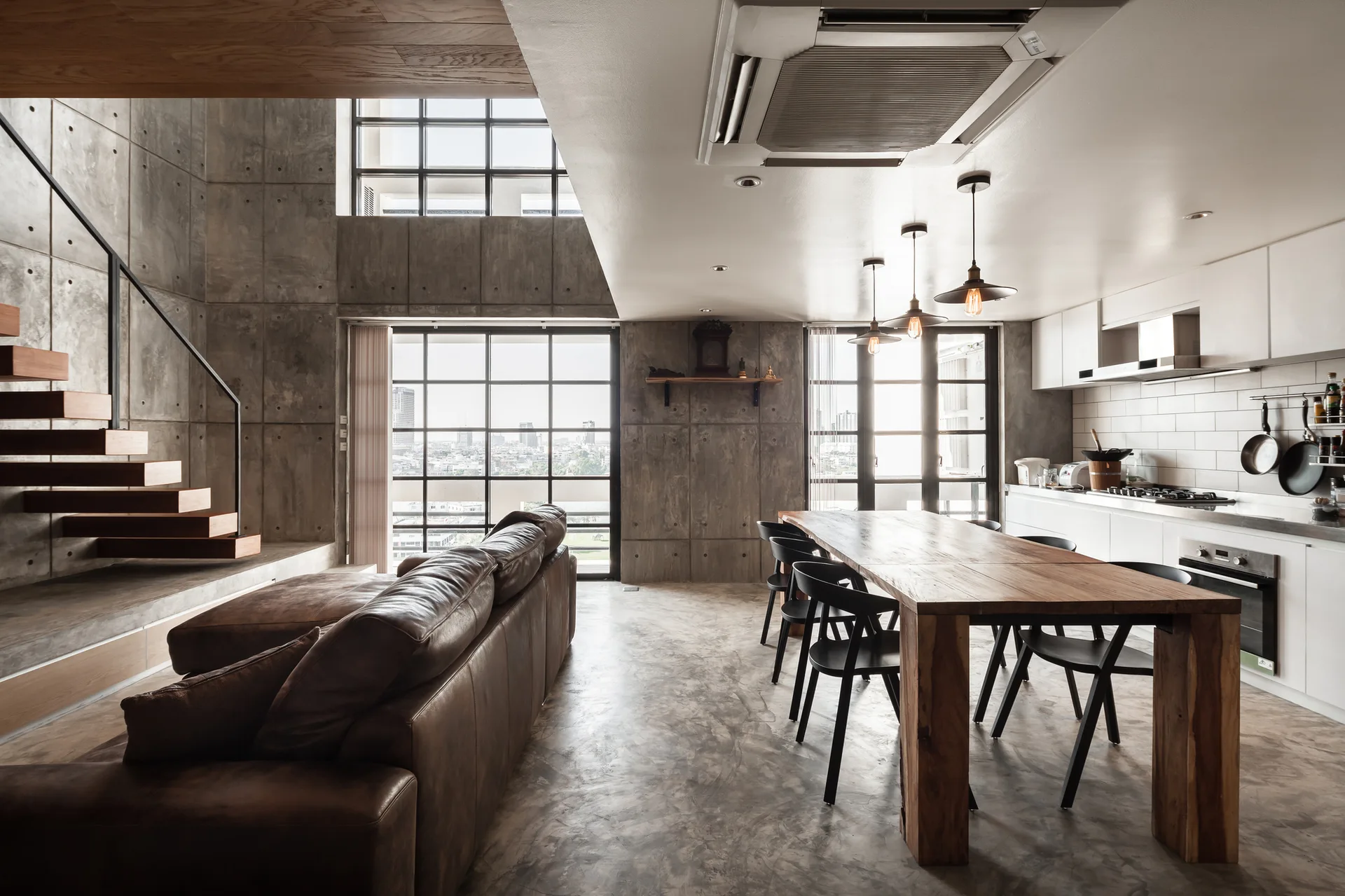 An open-plan industrial loft with concrete walls, a floating wooden staircase, a brown leather sofa, and a long wooden dining table.