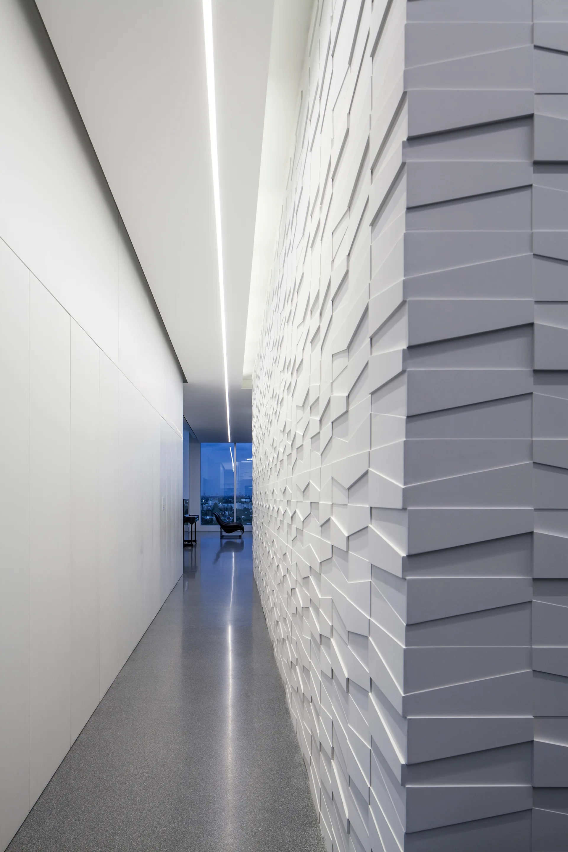 A long minimalist hallway features a white geometric relief wall, a continuous recessed linear light strip in the ceiling, and smooth grey flooring.