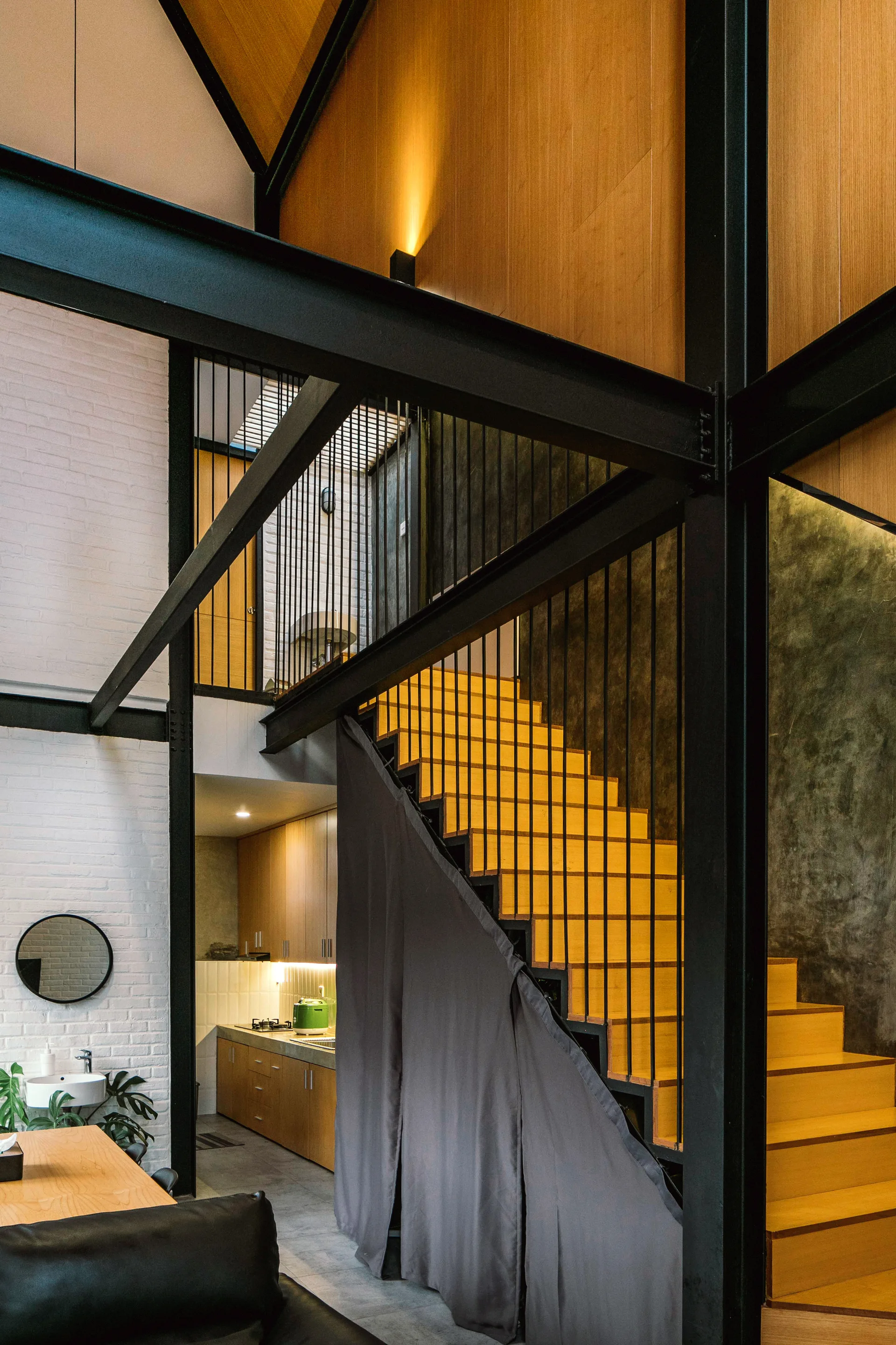 An industrial loft space with a wooden staircase, black structural steel beams, a white brick wall, and a dark curtain partition.