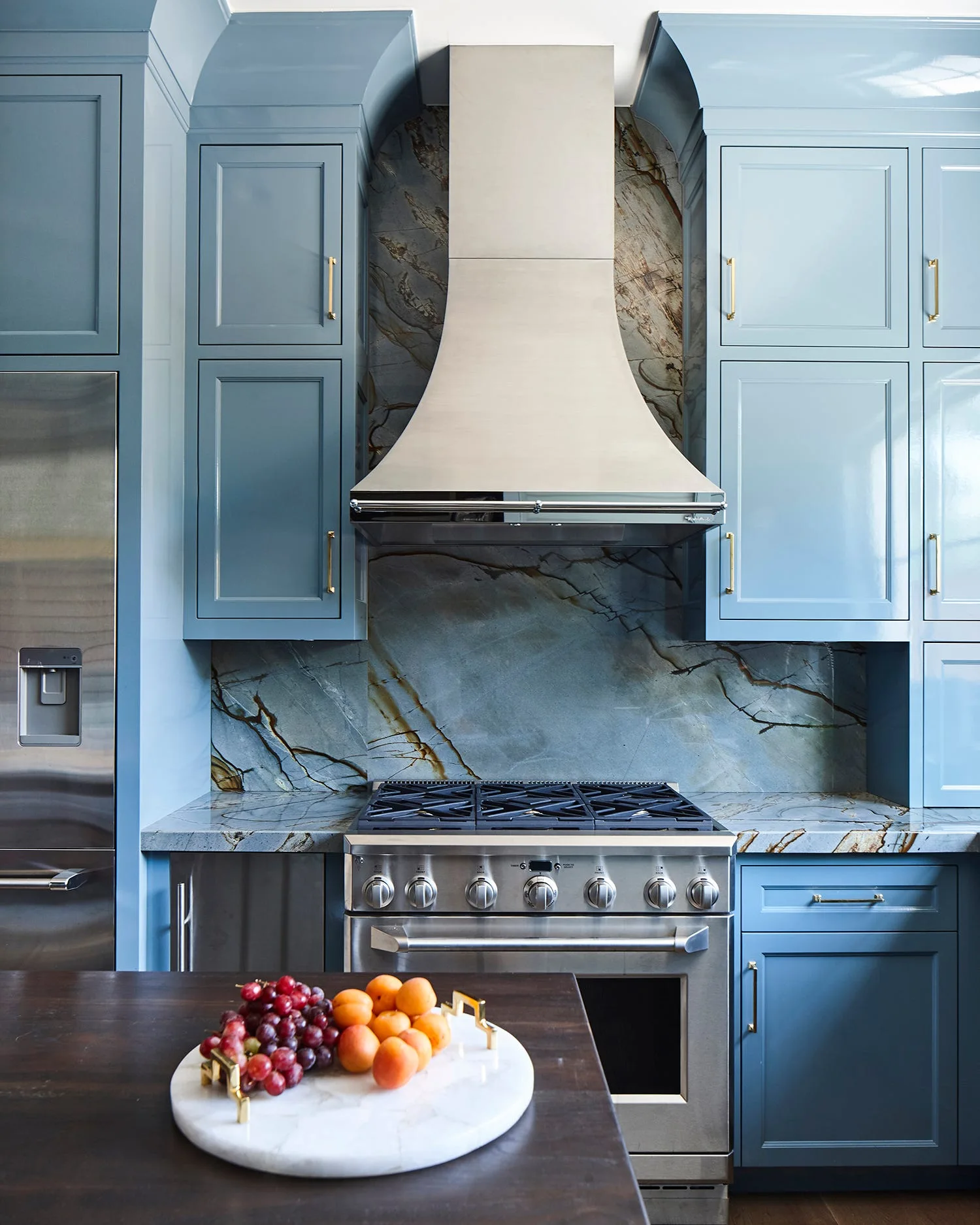 A kitchen featuring glossy blue cabinets with gold handles, a stainless steel gas range and hood, and a heavily veined stone slab backsplash.