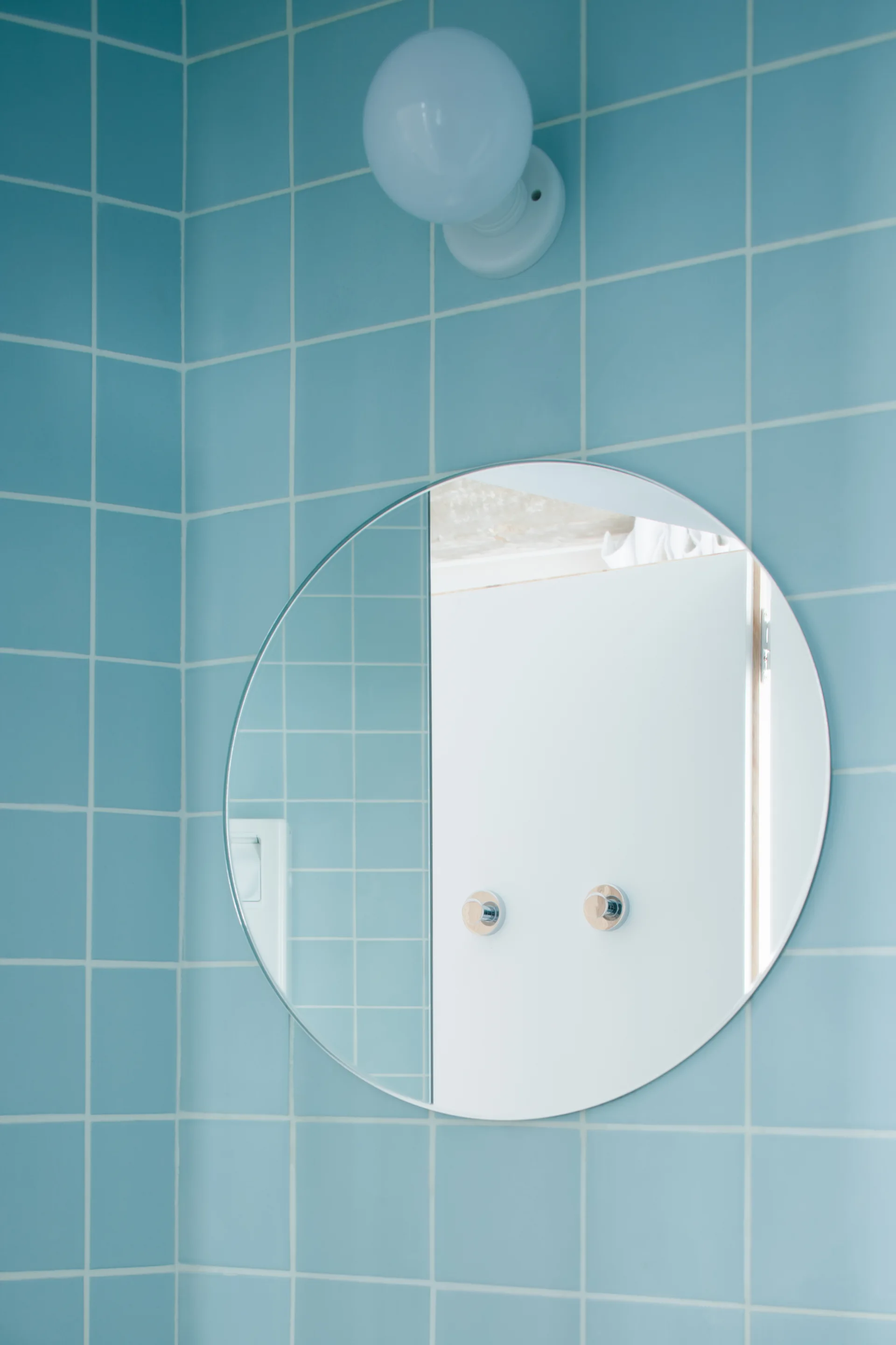 A glossy light blue tiled wall features a large frameless circular mirror with a white globe sconce mounted directly above.