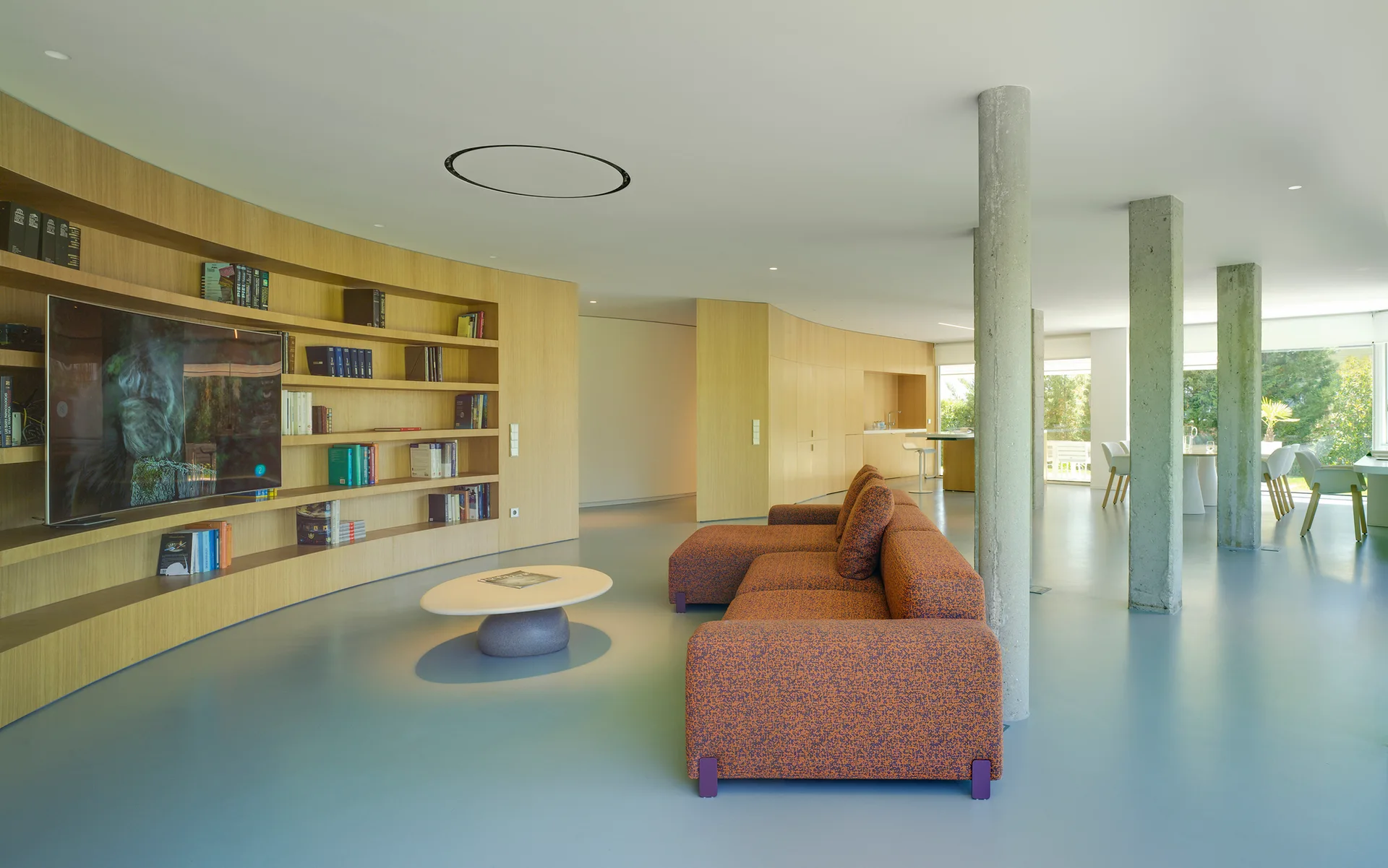 A minimalist living room features an orange textured sofa, raw concrete columns, and integrated light wood shelving against a smooth grey floor.