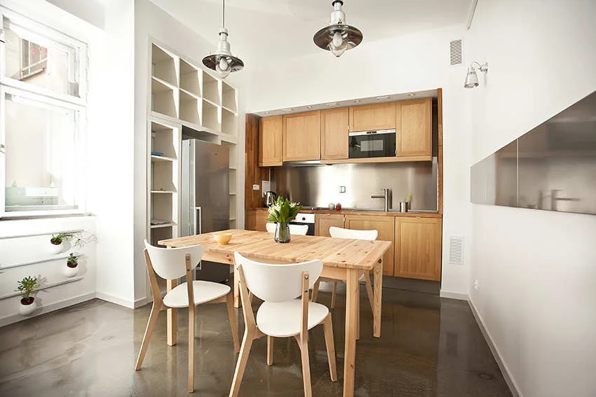 A Scandinavian kitchen features a wooden dining table, white chairs, natural wood cabinets, and built-in cubby shelving over a shiny concrete floor.