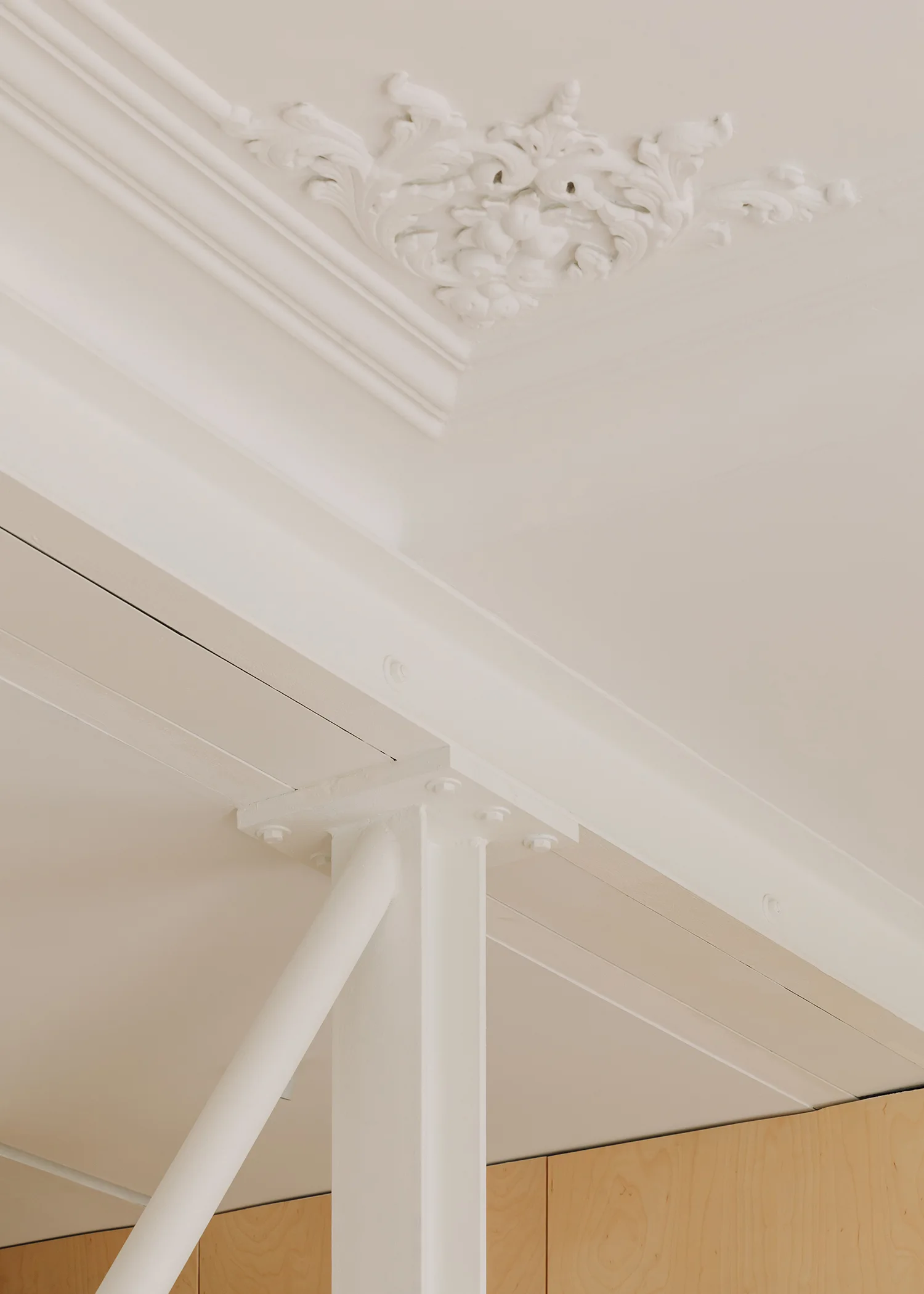 A white painted metal structural column and horizontal beam meet a white ceiling decorated with an ornate floral plaster medallion and crown molding.