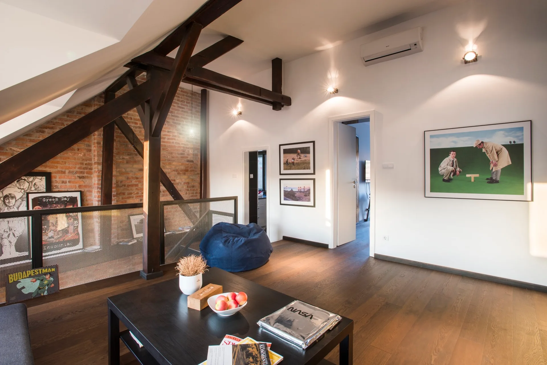 An attic living room featuring dark wood flooring, heavy exposed ceiling beams, a red brick accent wall, and a low black coffee table.