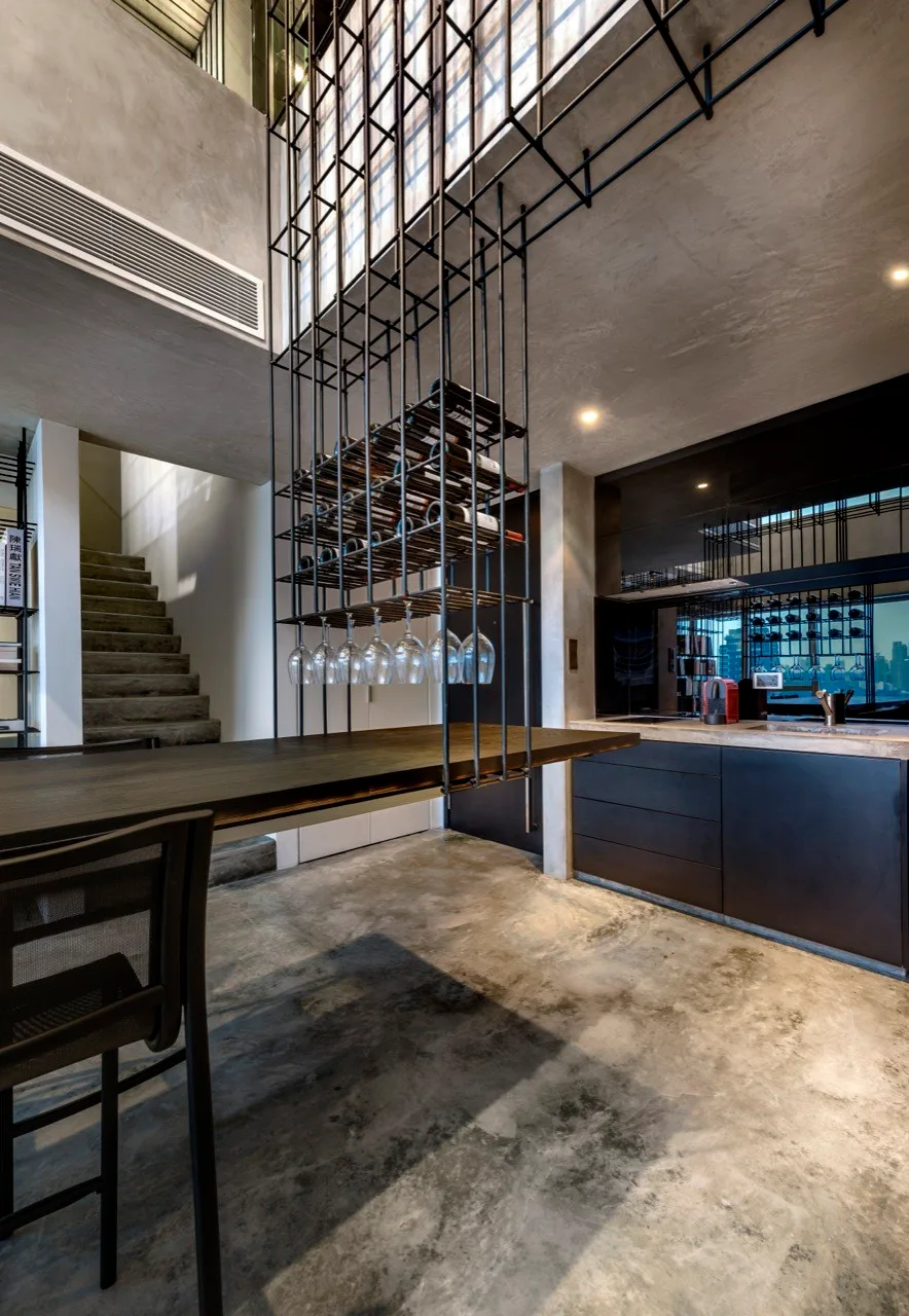 A modern industrial kitchen features black cabinetry, a textured concrete floor, and a suspended black metal wine rack above a dark wood table.