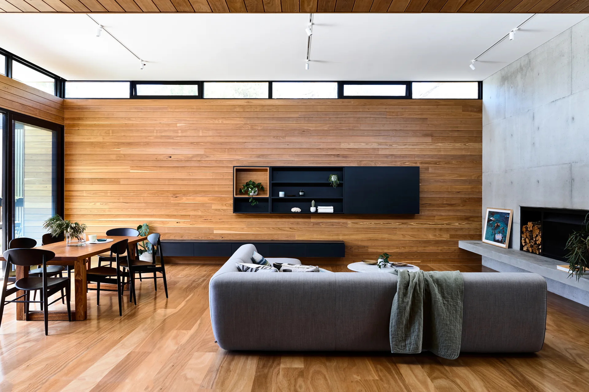 A modern open living room with a grey sofa, wooden dining table, warm timber paneled wall, and raw concrete fireplace wall.