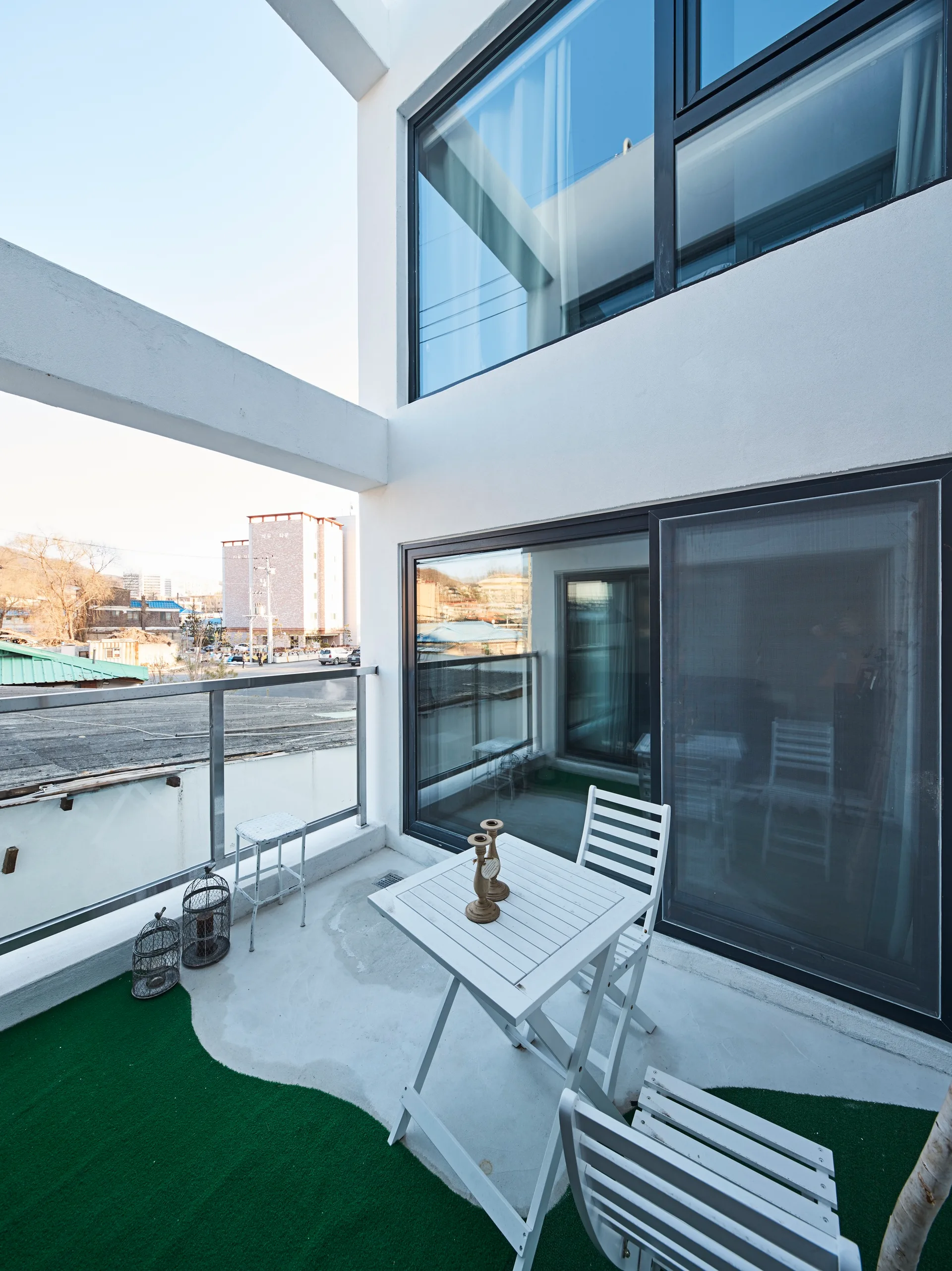 A concrete balcony features a white wooden bistro table and chairs, a curved patch of green artificial turf, and a glass railing.