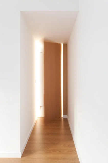 A minimalist white hallway with wood flooring features a wooden door accented by a warm, vertical integrated light glowing from the frame.