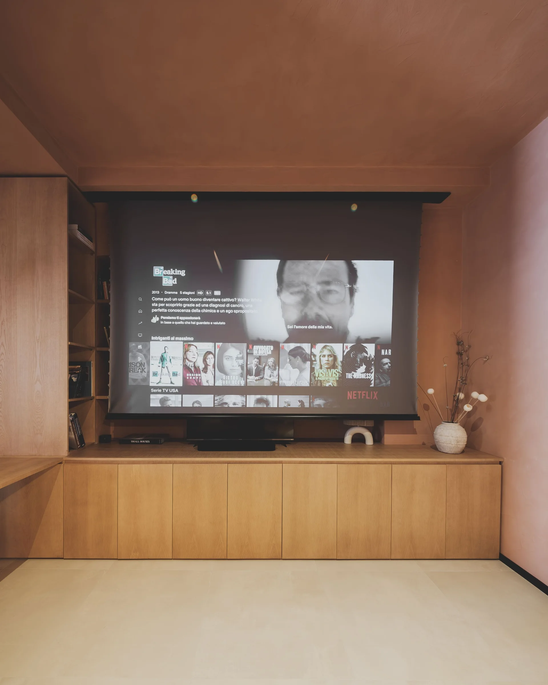 A home theater room features a projector screen mounted over low light wood cabinets against matte dusty pink walls and a matching ceiling.