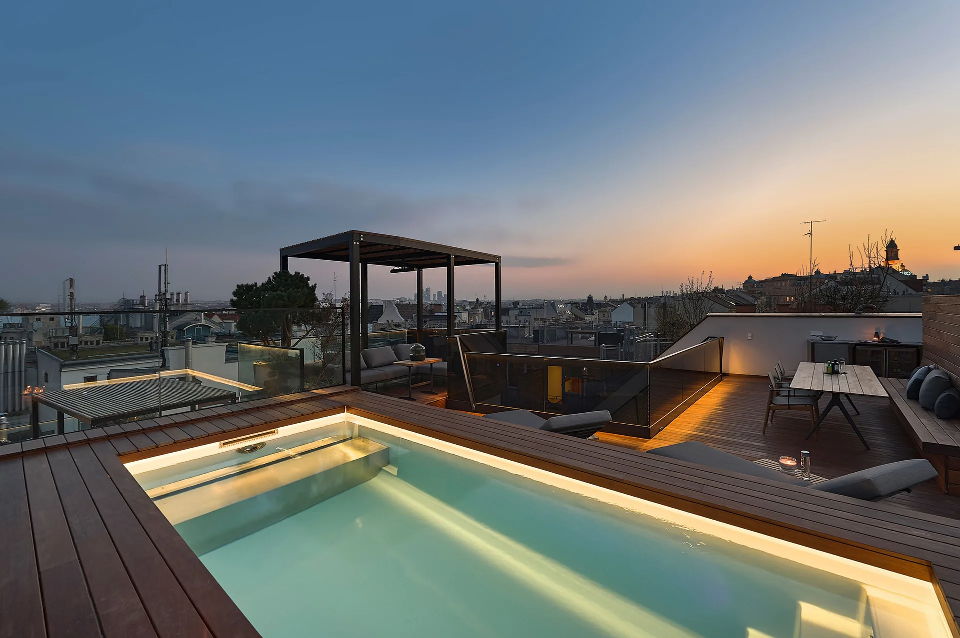 A wooden rooftop deck at dusk features an illuminated swimming pool, a dining table, and a shaded lounge area overlooking a city skyline.