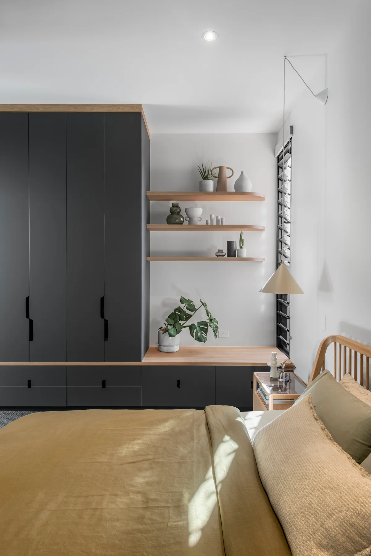 A modern bedroom featuring a dark grey built-in closet, floating wood shelves with pottery, and a light wood bed with warm yellow linens.