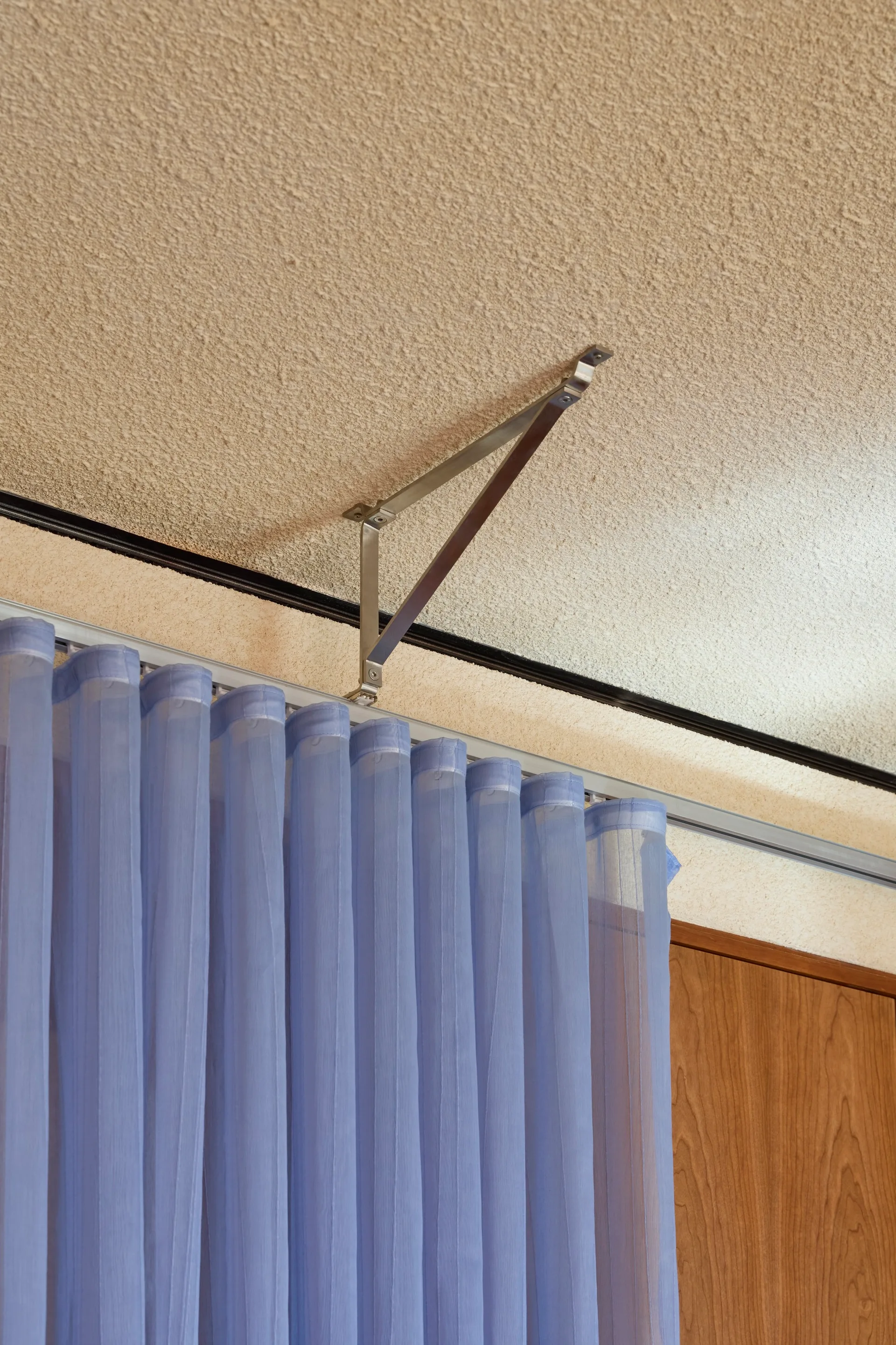 Close-up of a sheer blue curtain hanging from a silver metal track and bracket attached to a textured beige ceiling.