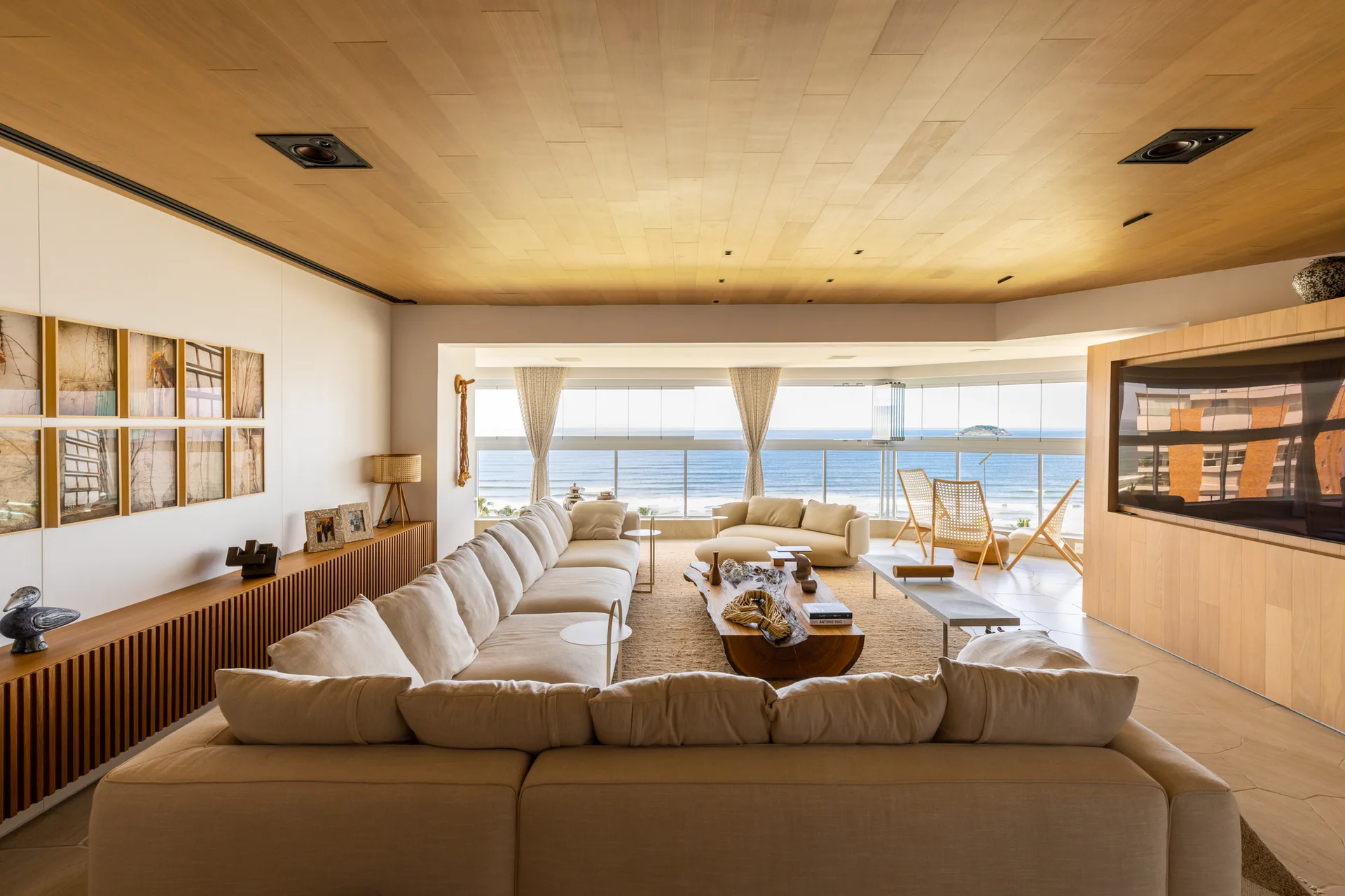 A spacious beachfront living room features a large beige sectional sofa, an organic wood coffee table, and panoramic ocean views through wide windows.