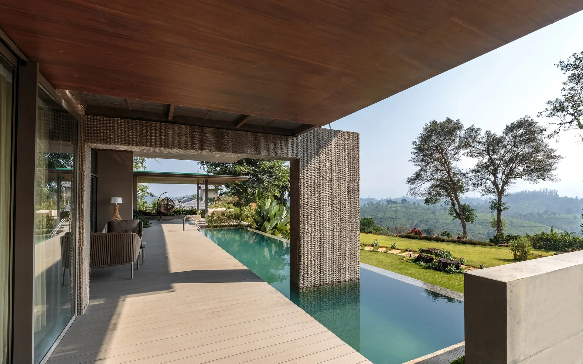 An outdoor patio deck features a blue infinity pool, a textured stone pillar, and a dark wood ceiling overhang facing green hills.