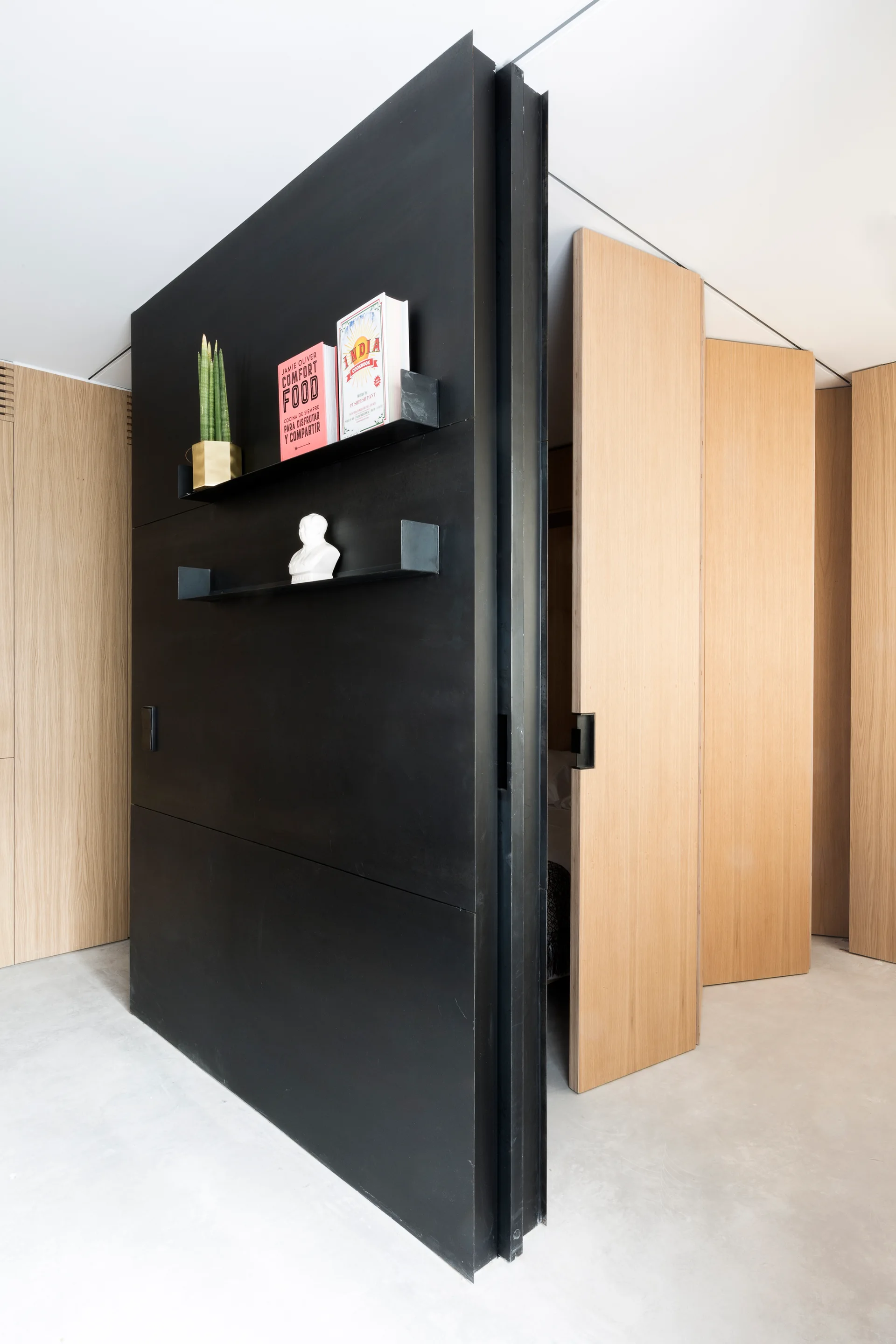 A matte black partition wall with thin metal display shelves stands next to light wood sliding panel doors on a light concrete floor.