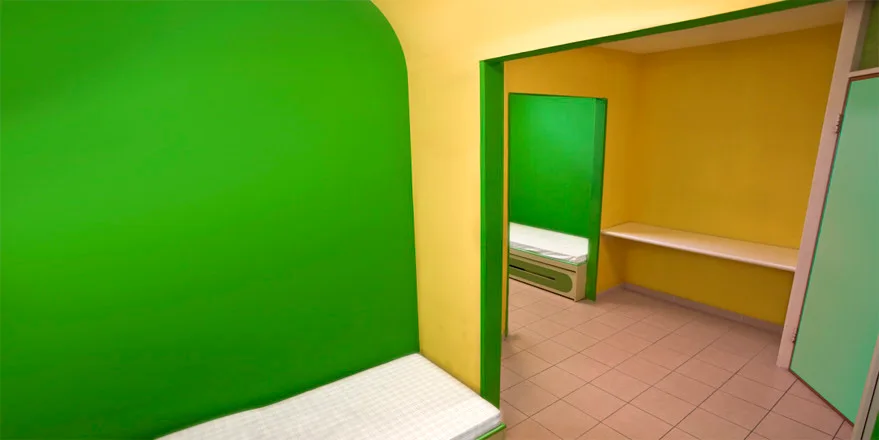 A simple room with bright yellow and green walls, featuring low-built platforms, a white shelf, and square-tiled flooring.