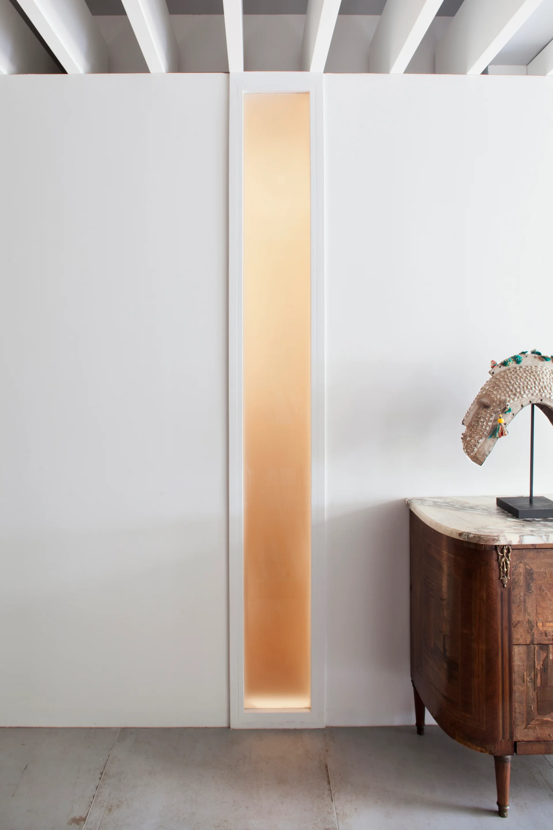 A wood console table with a marble top stands next to a white wall featuring an illuminated vertical glass panel under exposed ceiling beams.