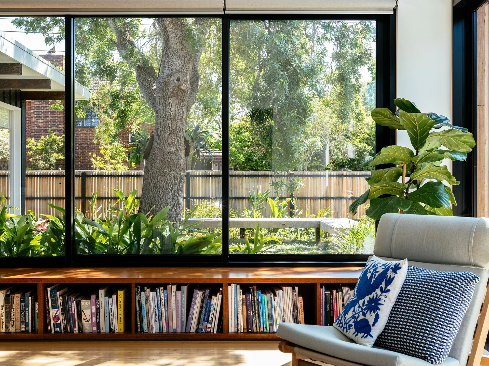 A sunlit reading nook features a low wooden bookshelf under black-framed windows overlooking a lush garden, with a modern armchair in the foreground.
