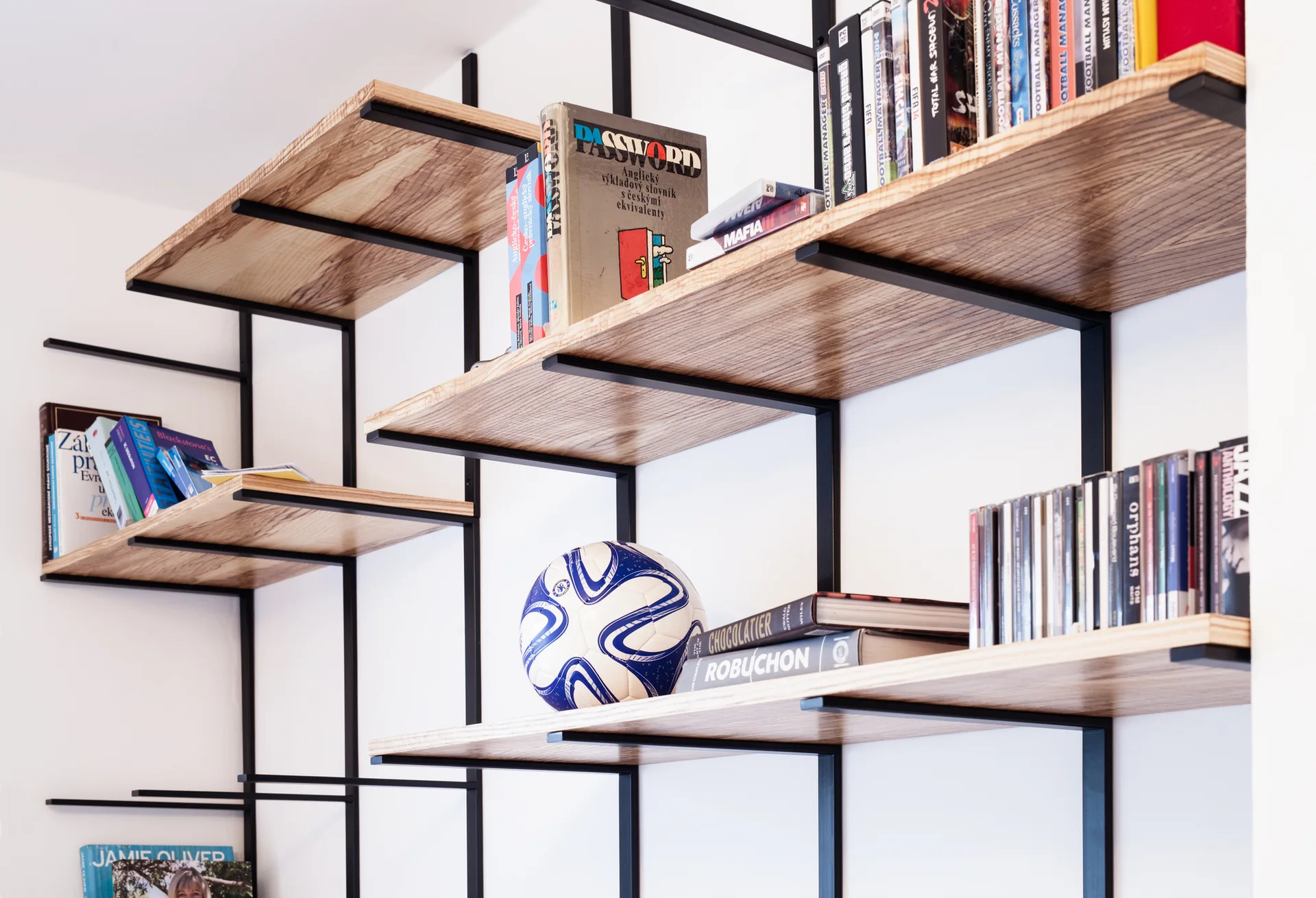 A wall-mounted shelving unit with light wood shelves and a black metal frame holds books, CD cases, and a blue and white soccer ball against a white wall.