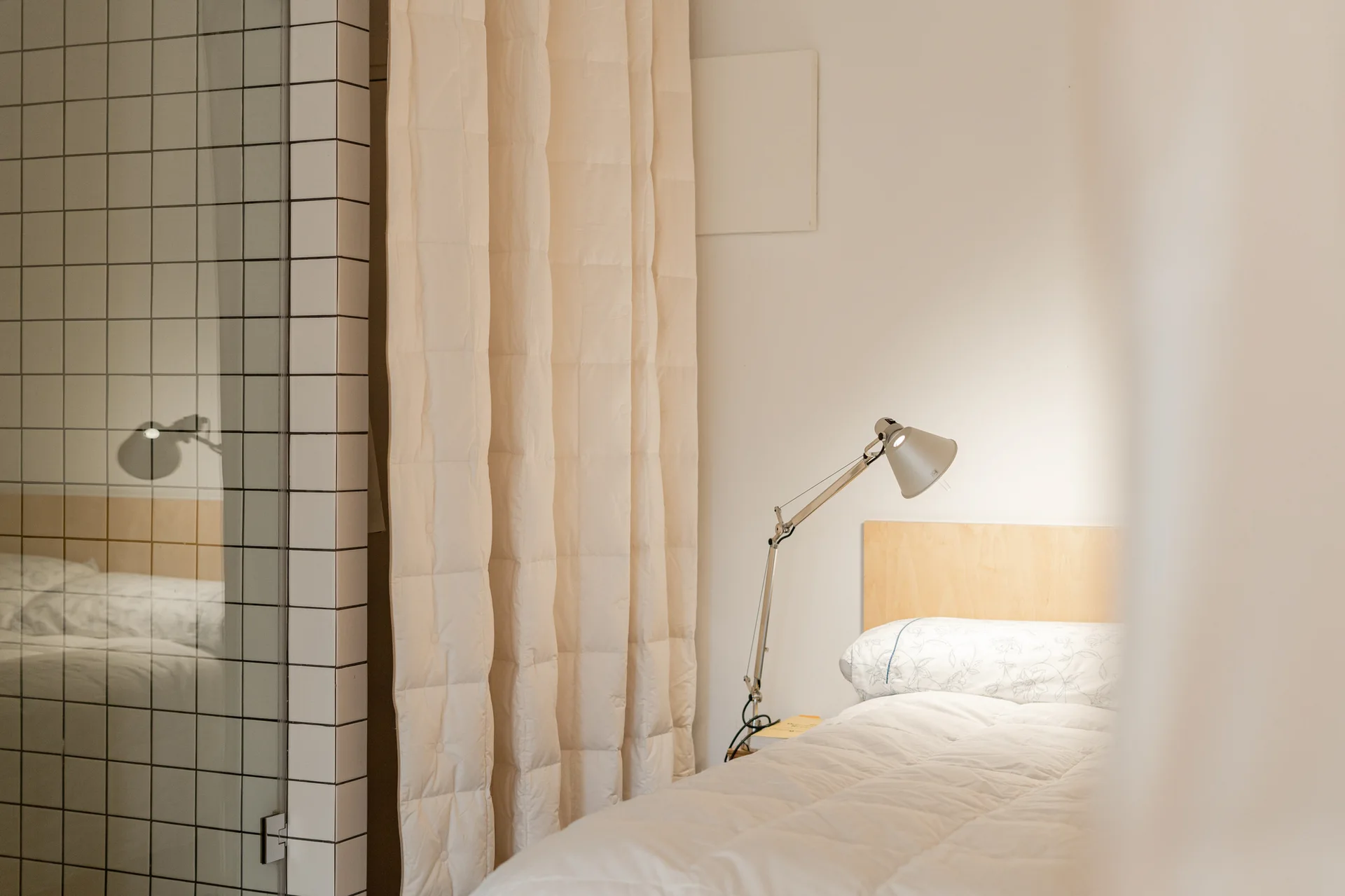 A minimalist bedroom featuring a bed with a light wood headboard, a white quilted comforter, an adjustable task lamp, and a white grid-tiled partition.