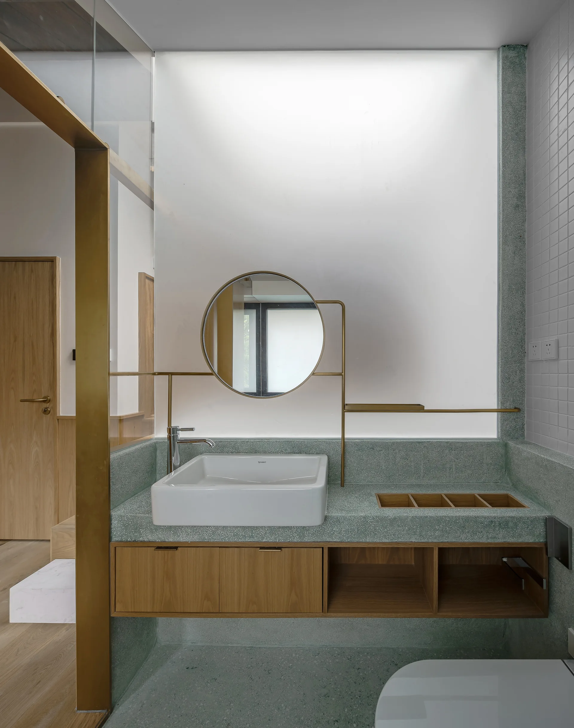 A modern bathroom features a sage green terrazzo vanity with an integrated sink, recessed wood storage cubbies, and a circular brass-framed mirror.