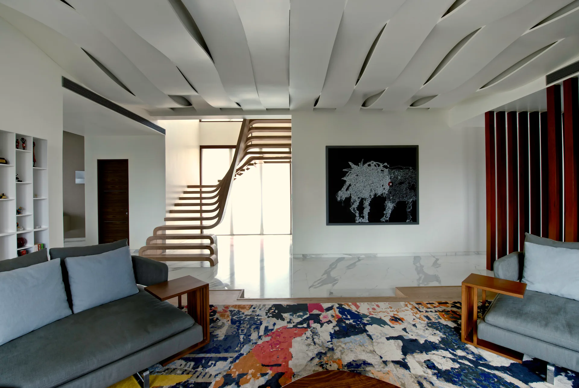 A contemporary living room features a sculptural wavy ceiling, gray sofas on an abstract rug, and a curved wooden staircase in the background.