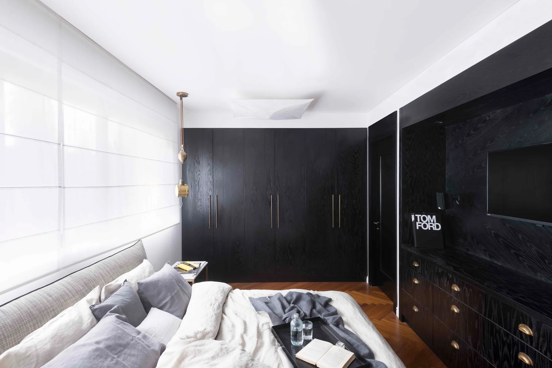 A modern bedroom features a large bed with linen sheets, dark wood built-in wardrobes, a matching dark dresser, and warm herringbone wood flooring.