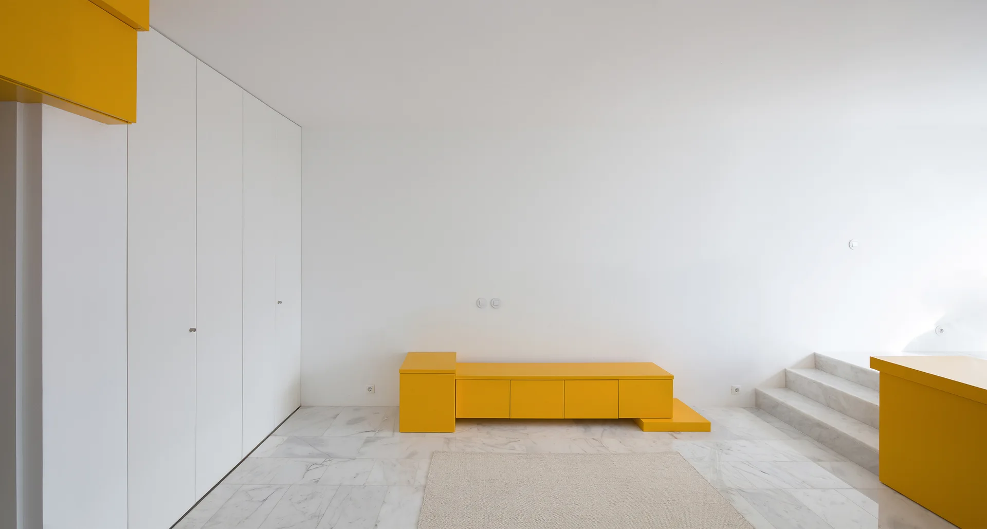A minimalist white interior features a bright yellow low cabinet, white built-in wardrobes, a beige rug on polished marble flooring, and marble steps.