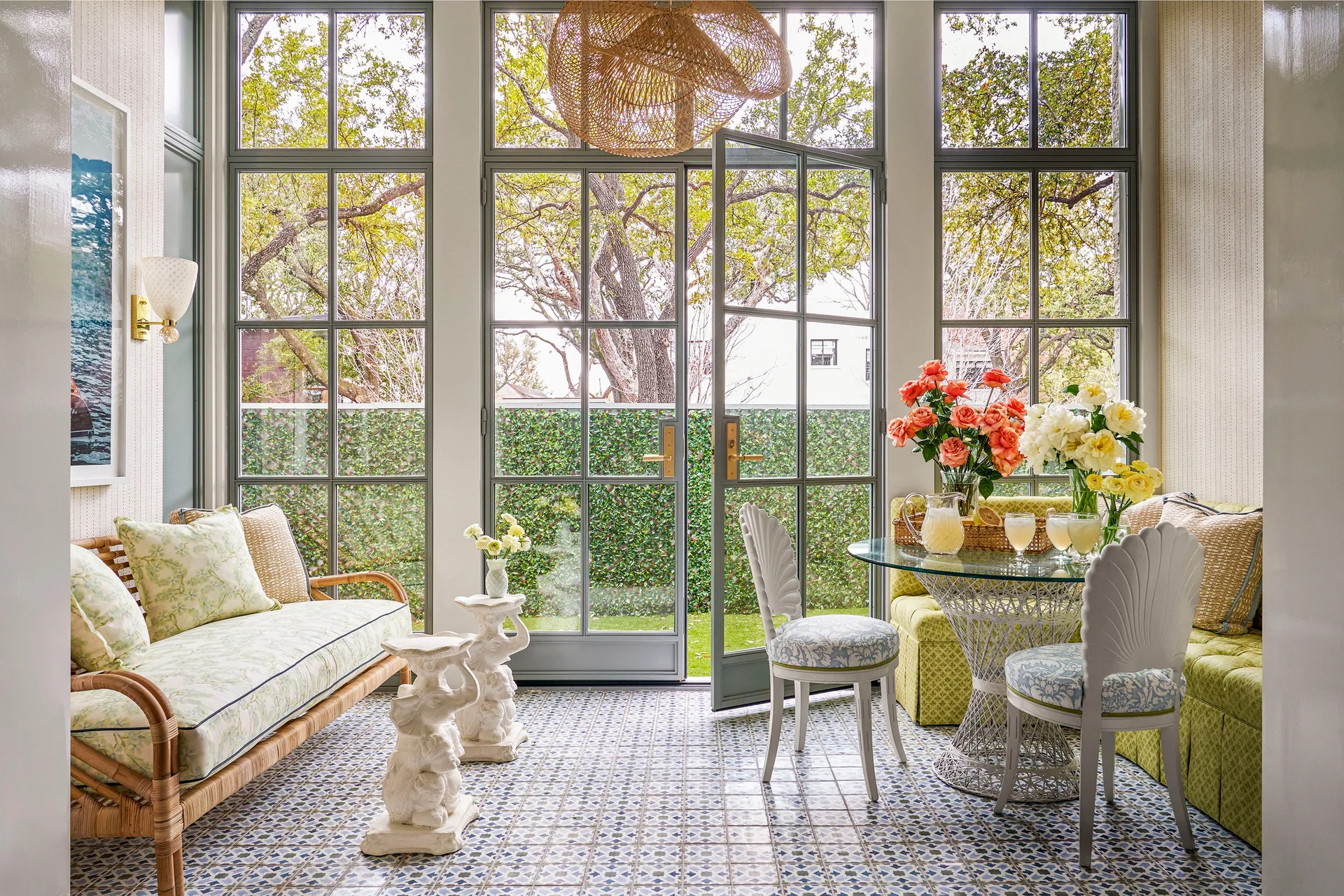 A sunroom features a rattan sofa, shell-back chairs around a glass table, patterned blue tile floor, and large metal-framed windows.