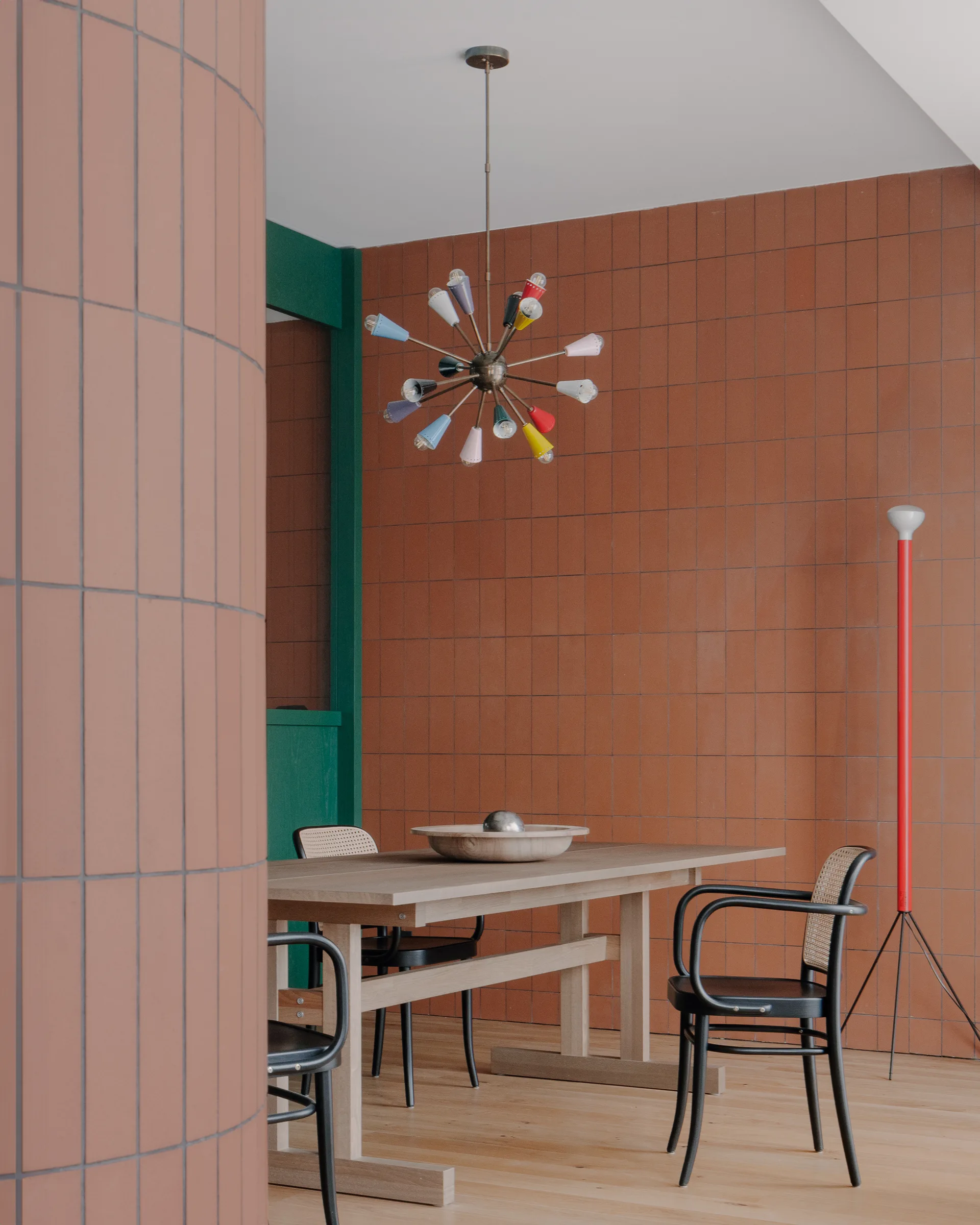A dining space features a light wood table and black caned chairs against a terracotta tiled grid wall with a colorful sputnik chandelier.