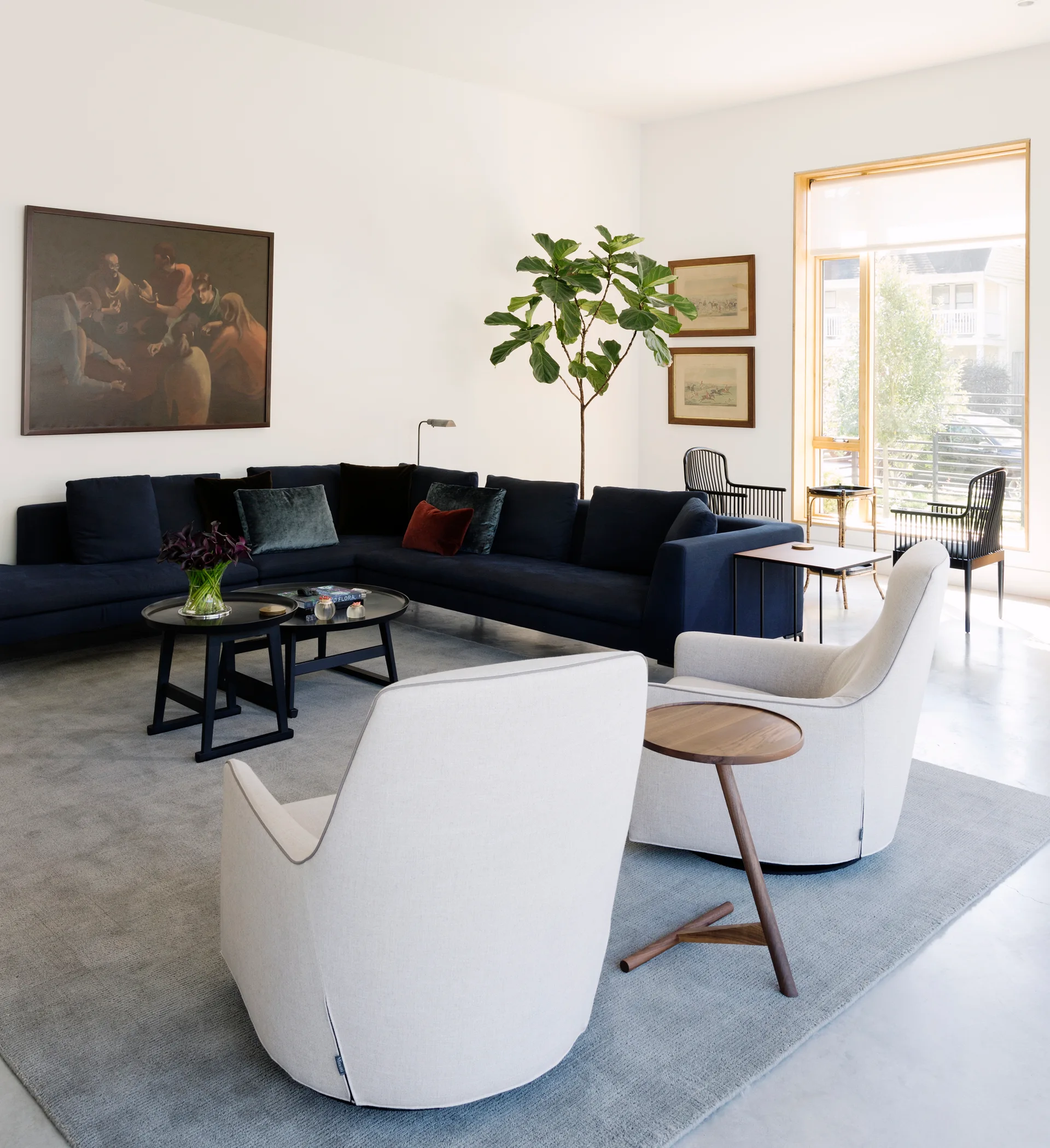 A modern living room features a dark navy sectional sofa, two light grey armchairs, nested black coffee tables, and a large wood-framed window.