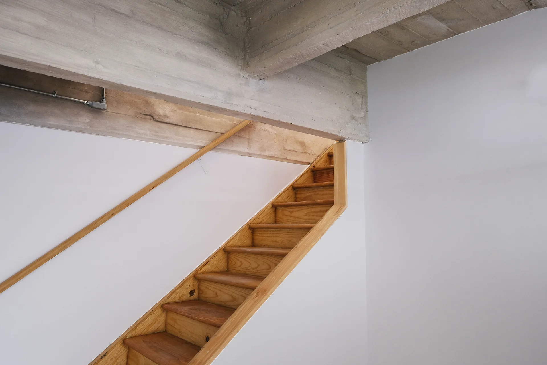 A wooden staircase with a slim matching handrail climbs a white wall beneath an exposed concrete ceiling beam.