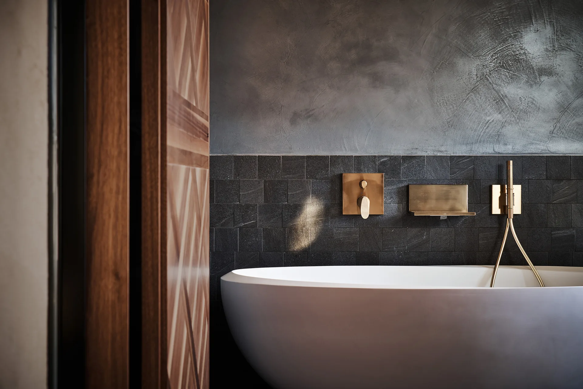 A white freestanding bathtub set against a wall with dark square stone tiles and a textured gray plaster upper section.