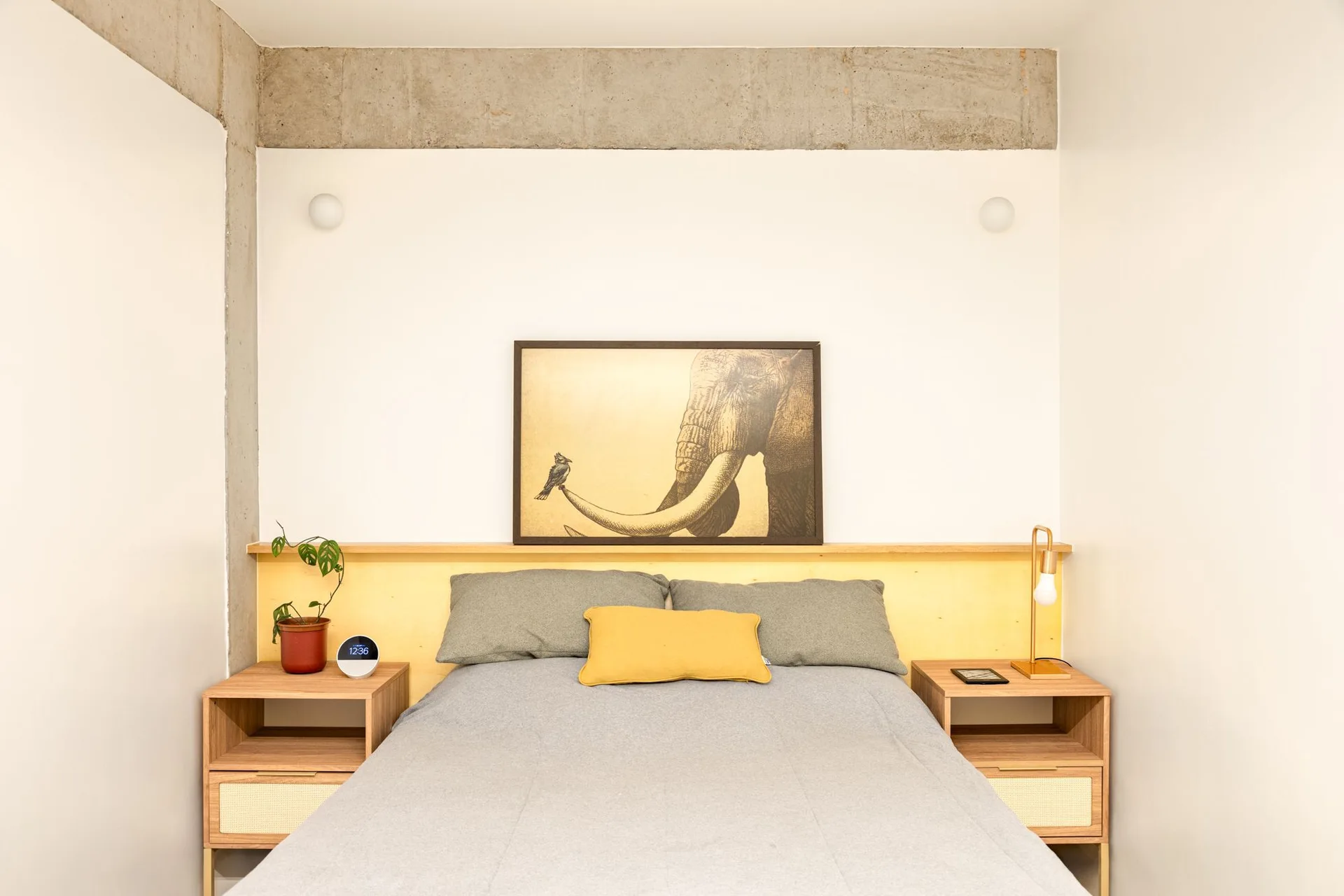 A bed with grey bedding sits between two wood nightstands under a yellow headboard panel, framed elephant artwork, and an exposed concrete beam.