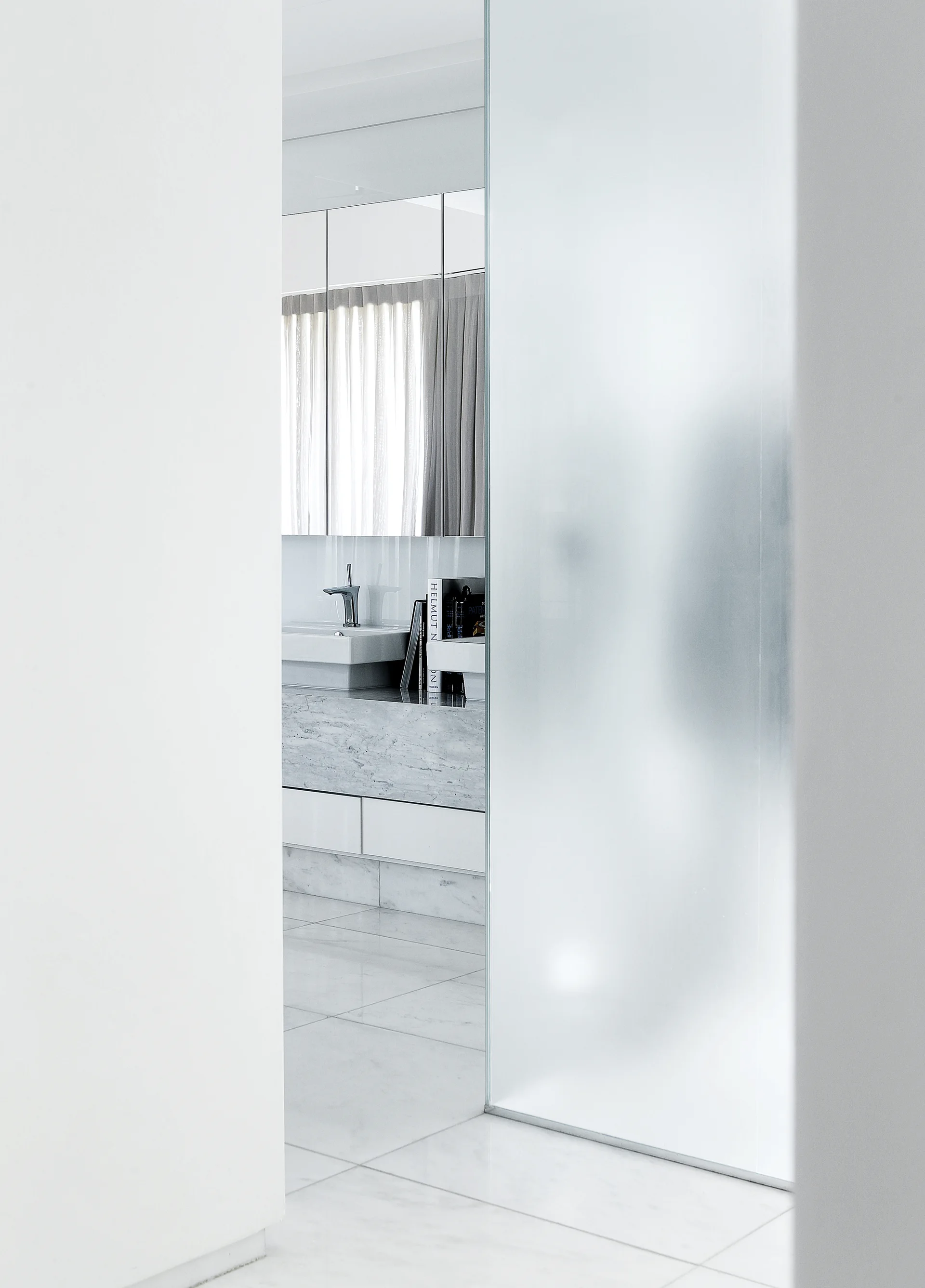 A view through a doorway into a white marble bathroom featuring a frosted glass partition, marble vanity, sink, and mirrored cabinets.