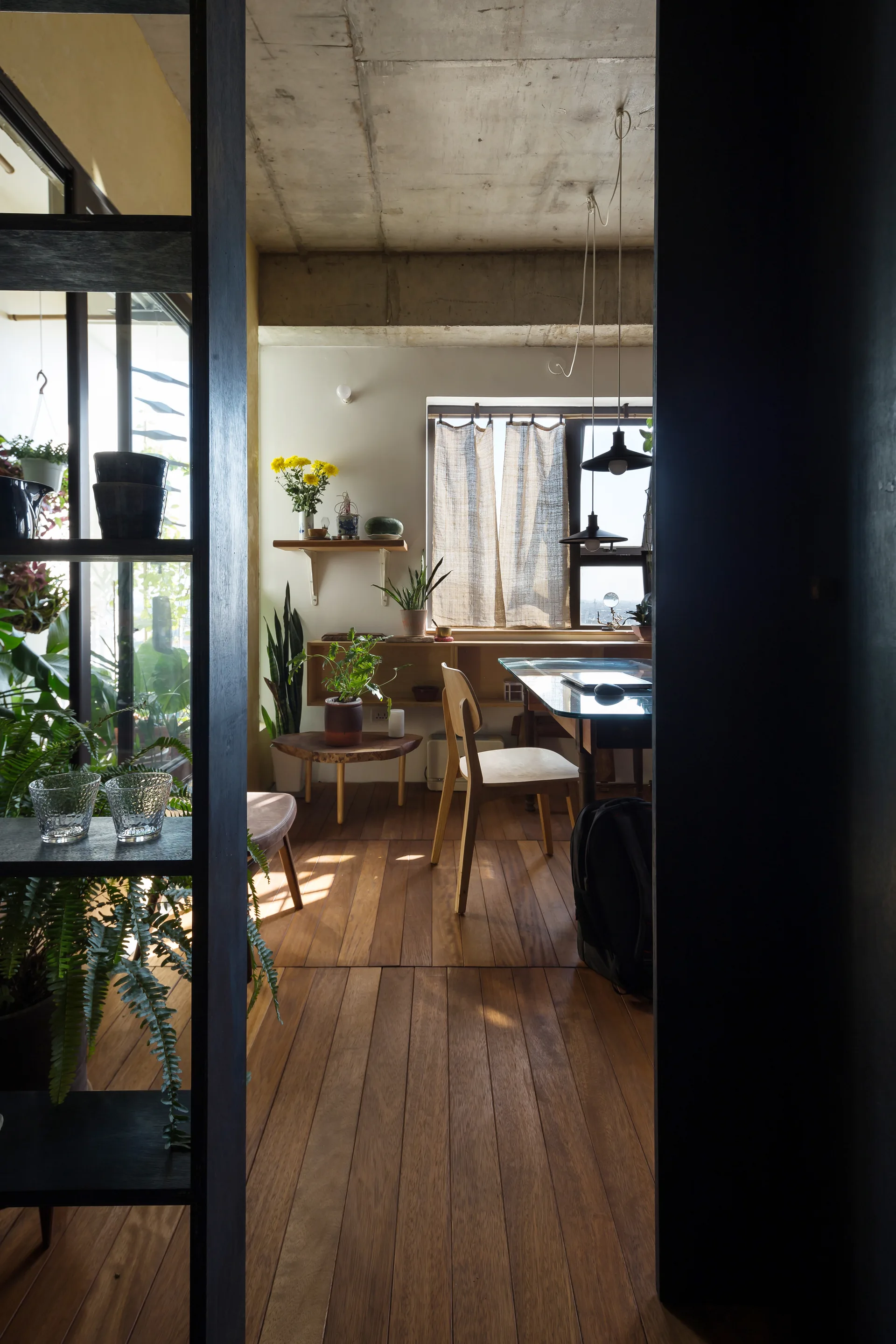 A home office viewed through a dark entryway features an exposed concrete ceiling, wood plank flooring, a wooden desk and chair, and indoor plants.