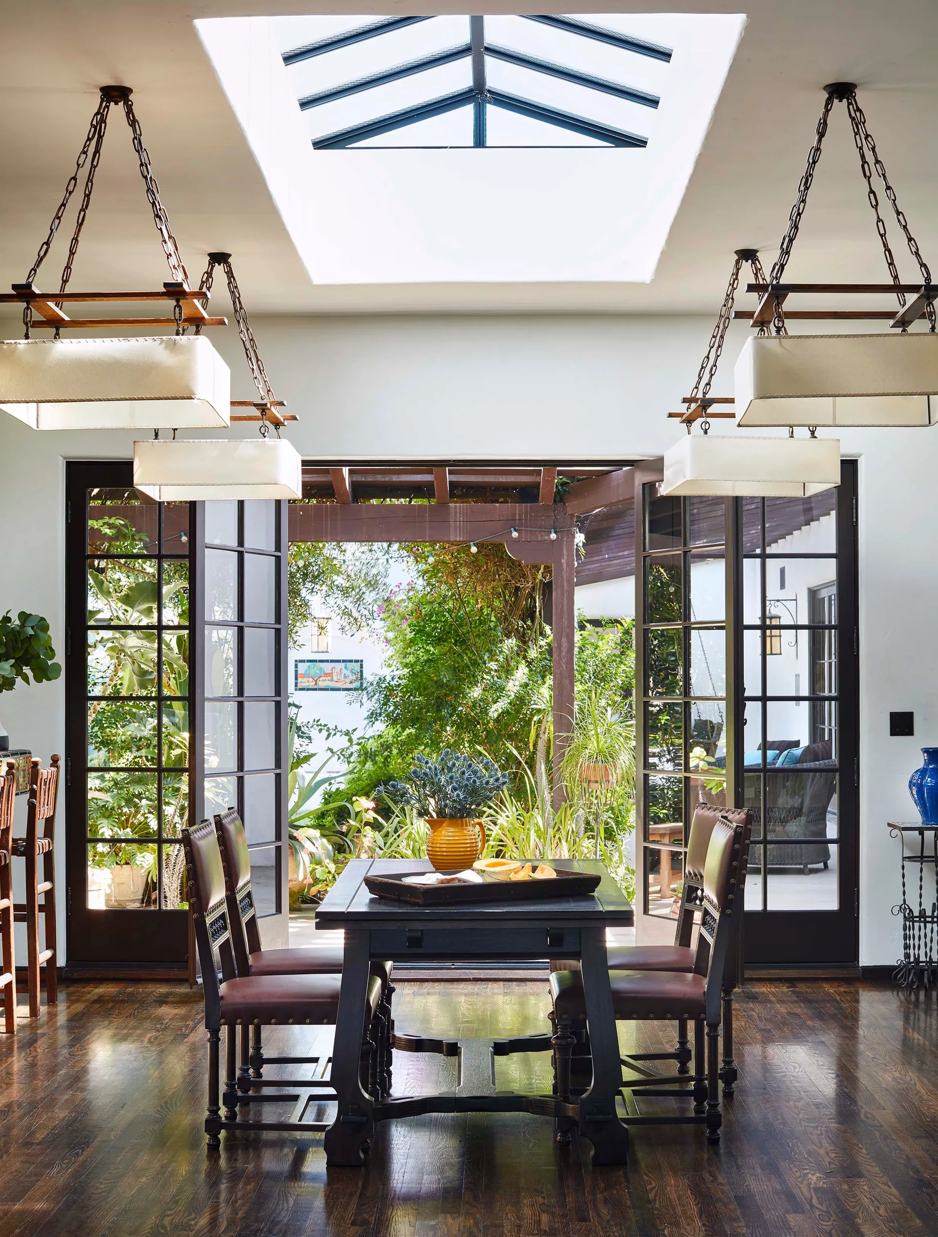 A dark wood dining set sits under a skylight and fabric pendant lights, with open French doors showing a lush courtyard garden outside.