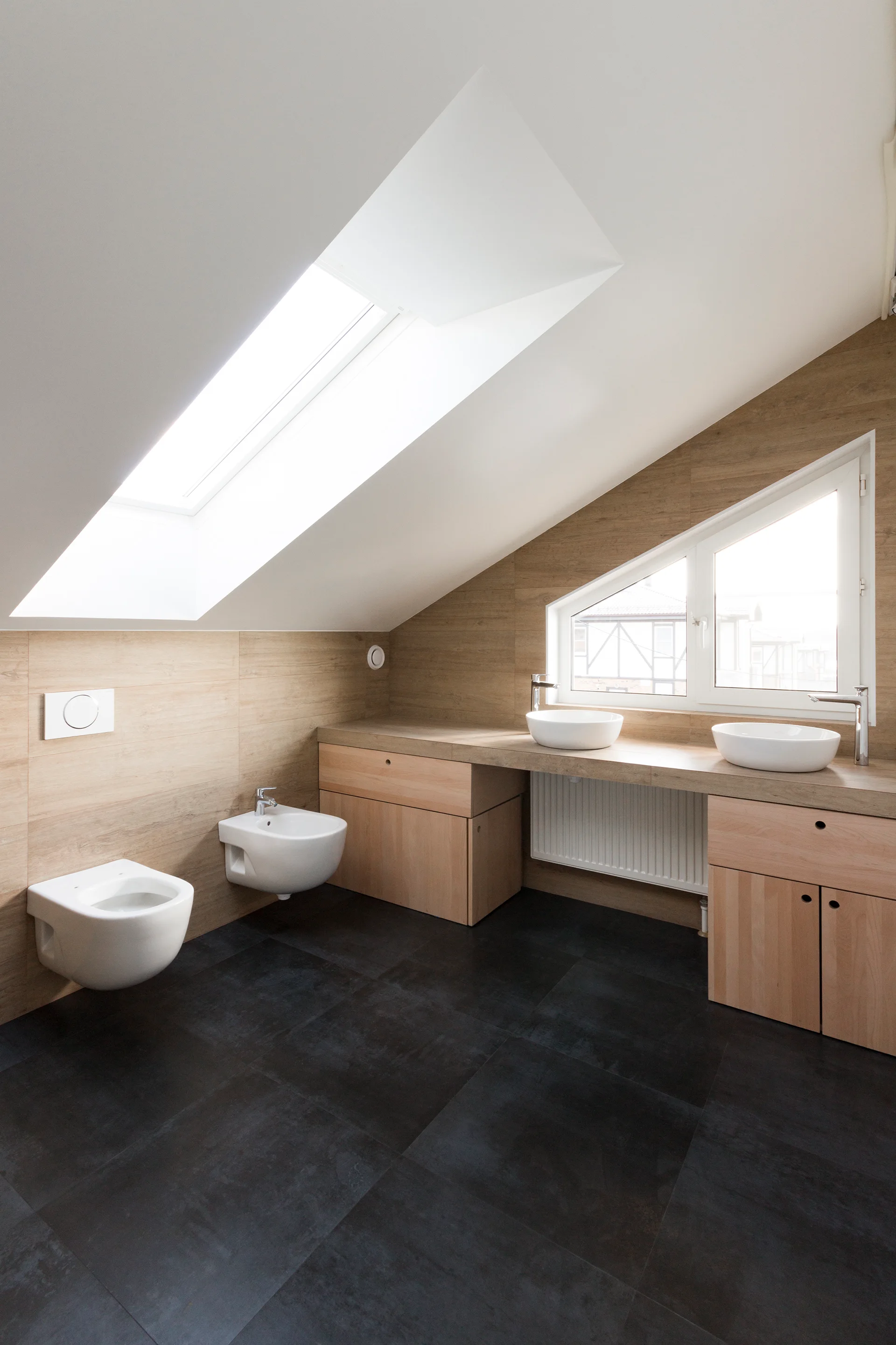 A minimalist attic bathroom featuring a sloped ceiling with a skylight, wood-paneled walls, a light wood vanity, and dark floor tiles.