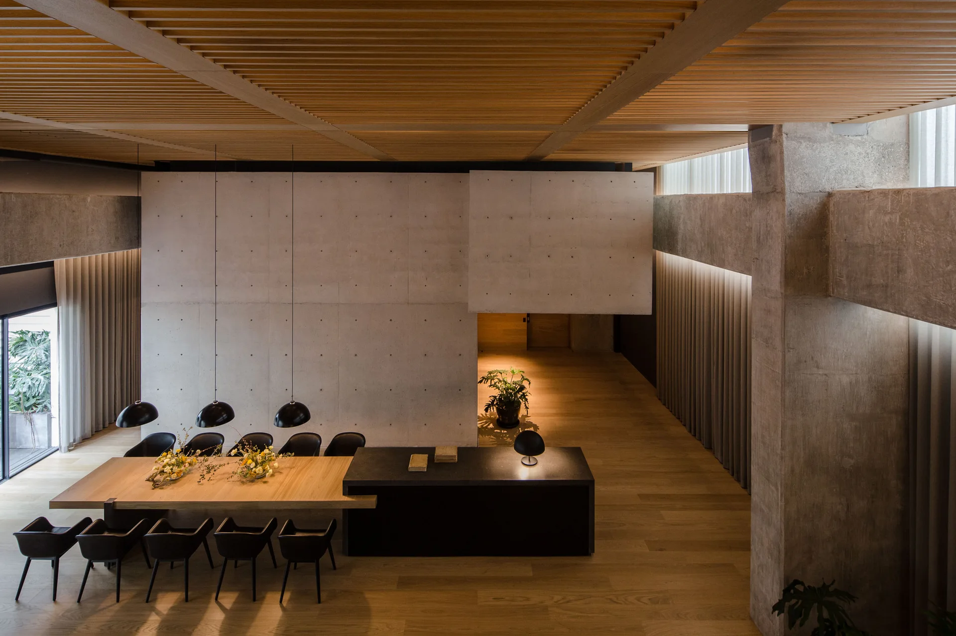 A modern dining area features a long wooden table integrated with a black island beneath a slatted wood ceiling and next to a concrete wall.